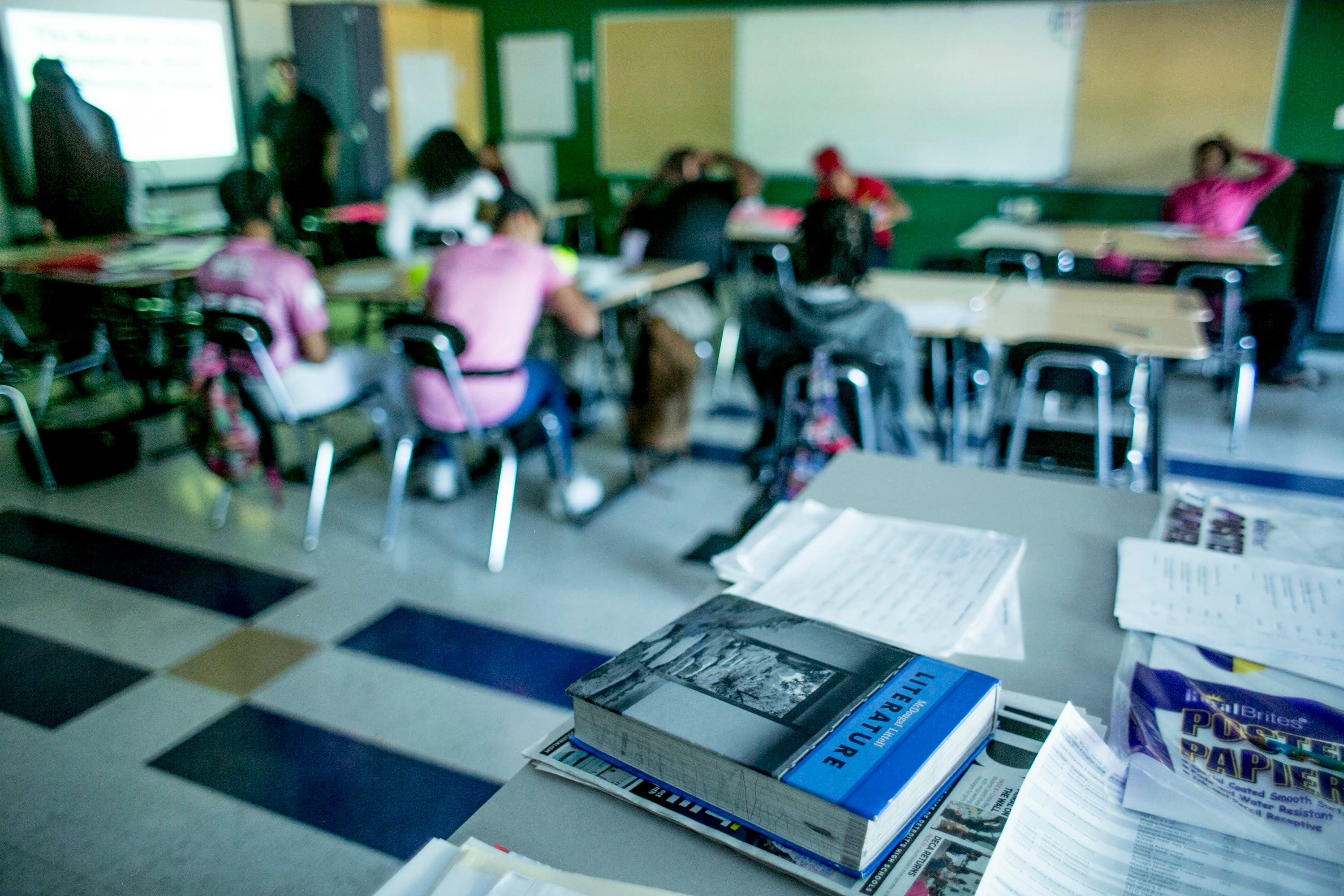 Textbooks are in the foreground with students seated at desks in the background.