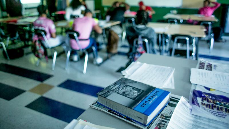 Textbooks are in the foreground with students seated at desks in the background.