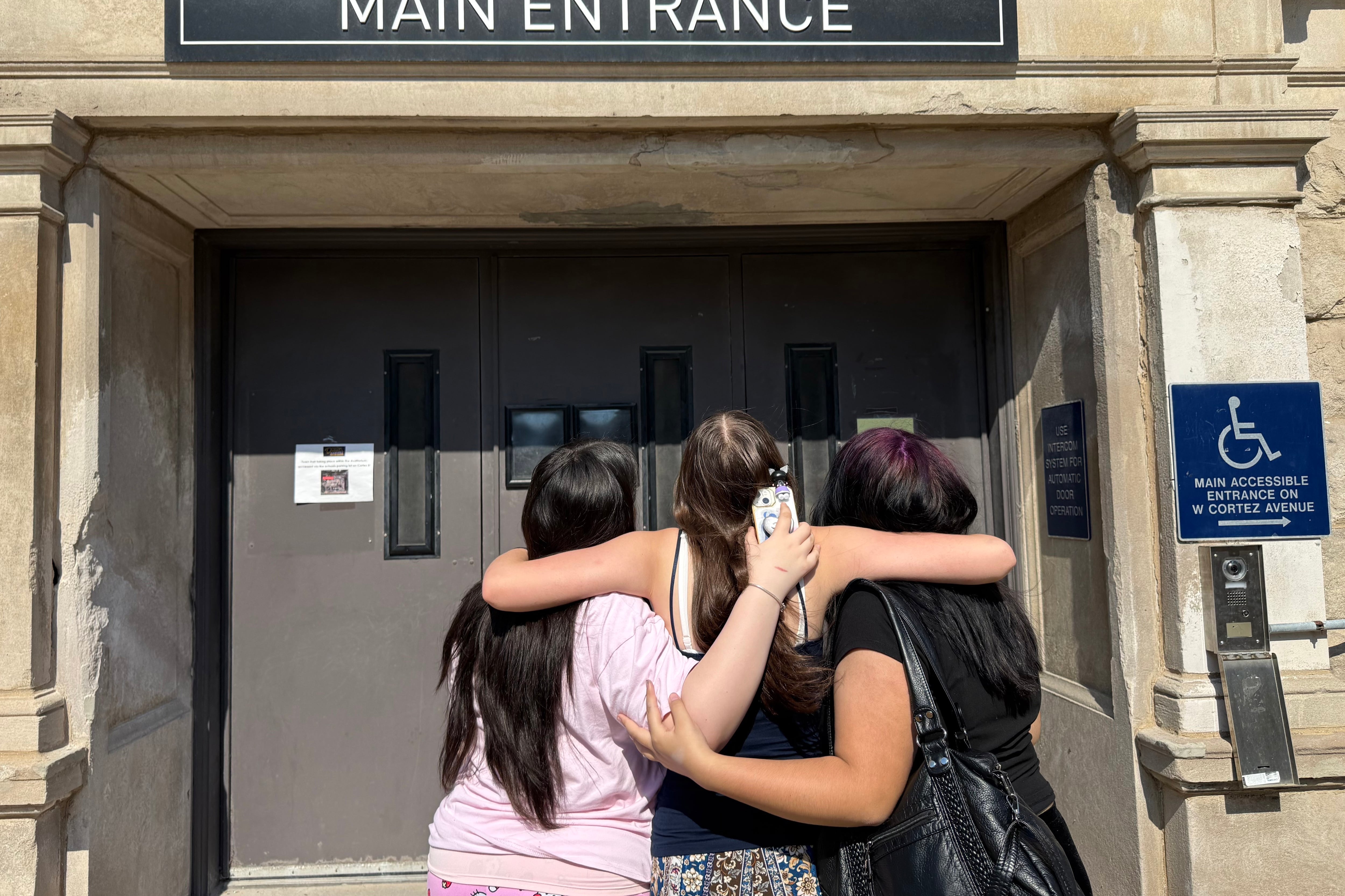 Three female students hug in front of a door that says Main Entrance3.