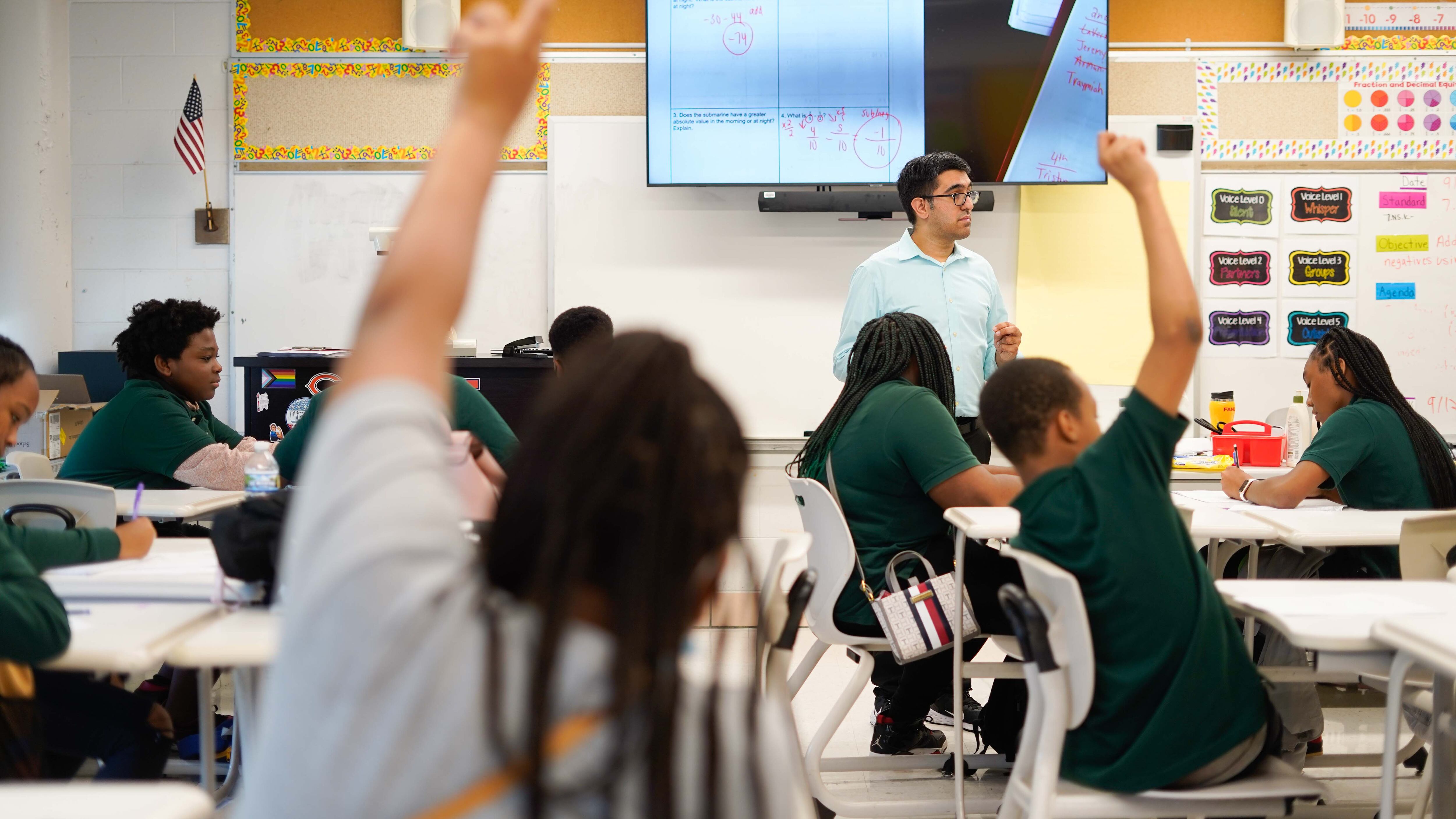 Two students raise their hands in class while a teacher wearing glasses and a blue shirt stand in the background looking at a classroom full of students.