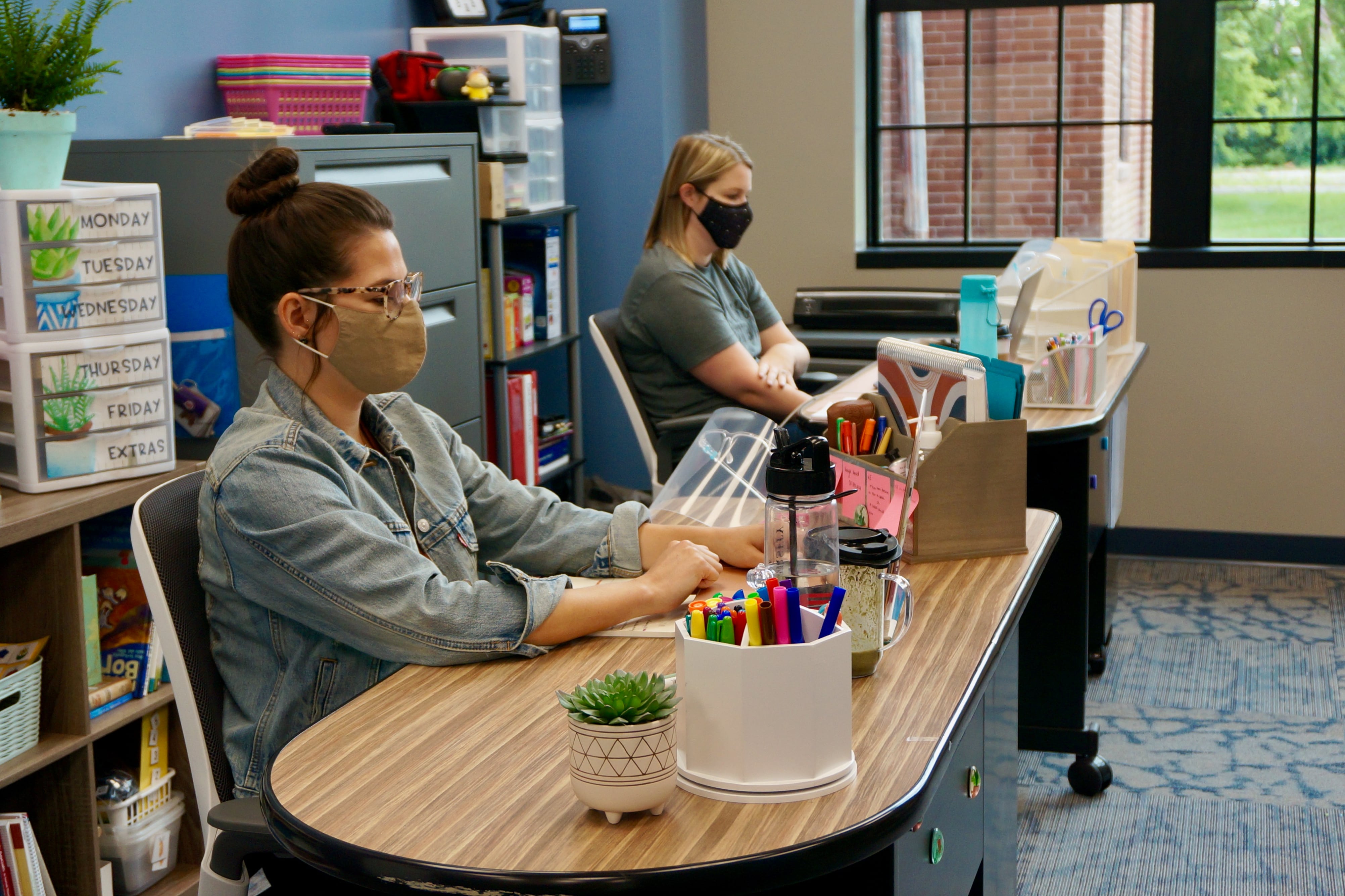 Two teachers sit in a classroom at laptops with masks on.