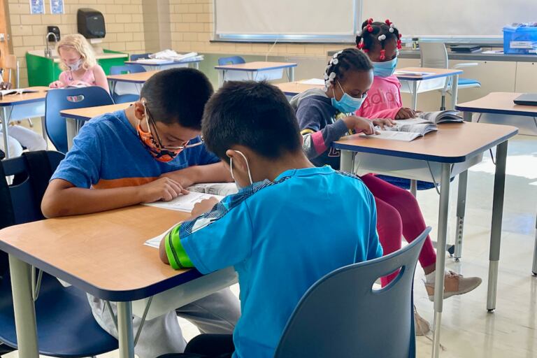Young students work at their desks in a classroom. The two boys in the foreground are both wearing blue shirts and protective masks.