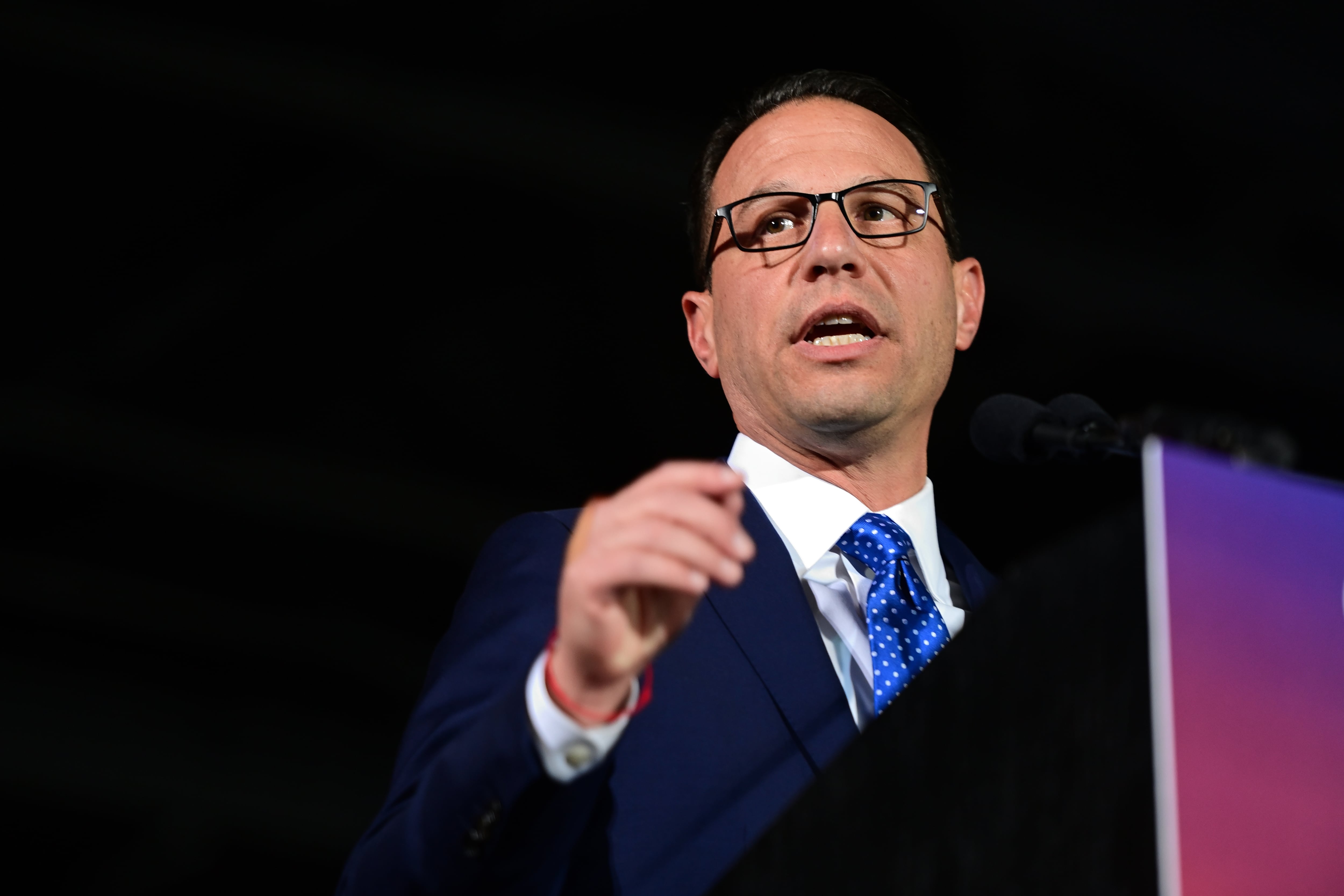 A man wearing glasses, a dark suit, and a blue tie speaks from a podium while raising his hand.