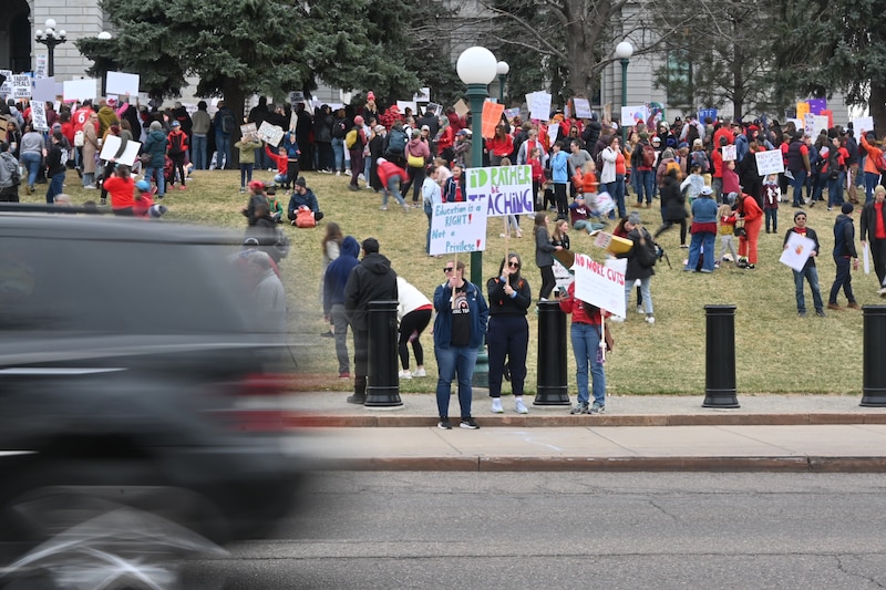 A group of adults stand outside of a large Capitol building, some holding protest signs while a car passes in front.