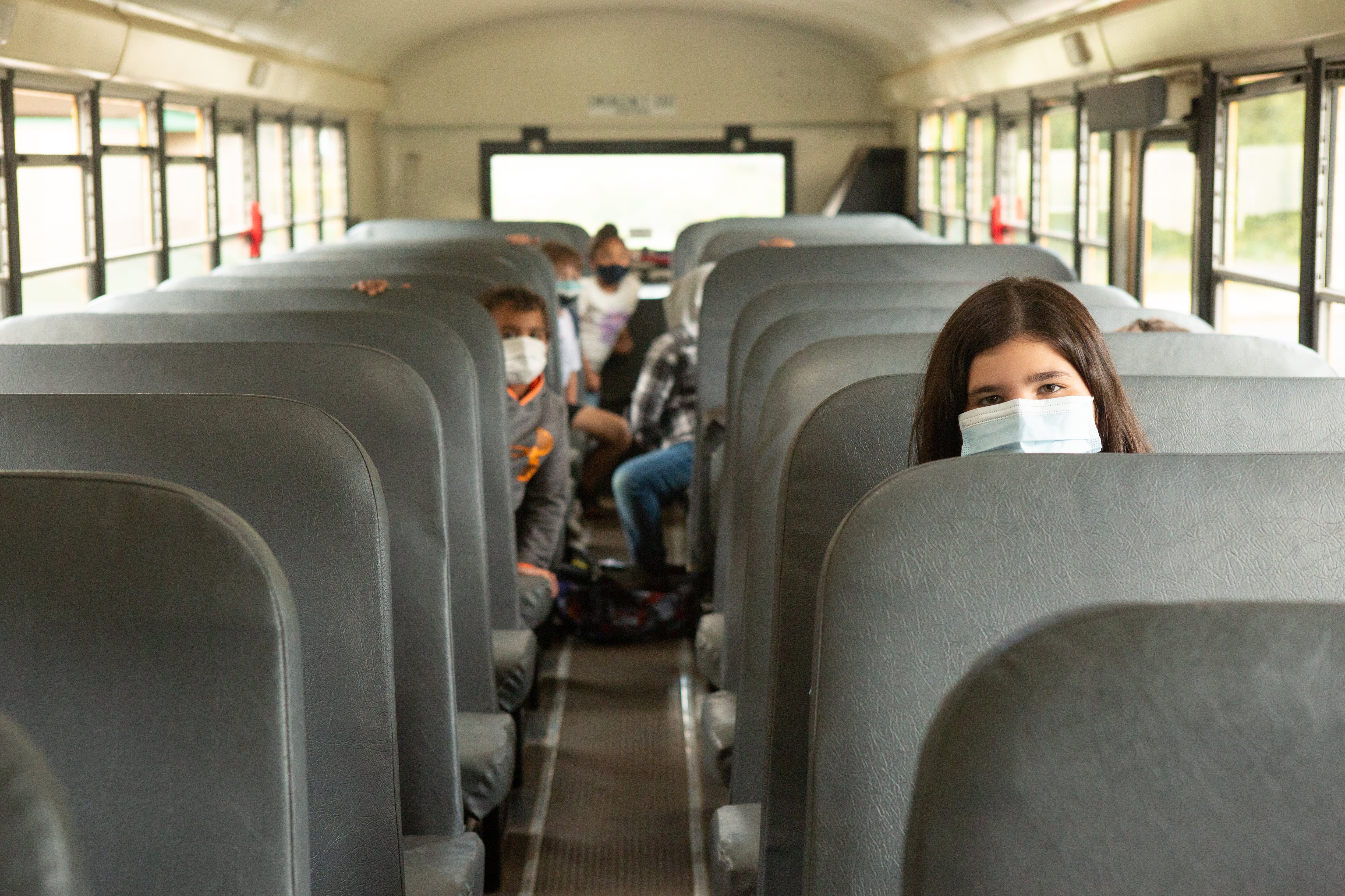 Several students, wearing blue face masks, sit on a bus with gray seats waiting as students wait to depart from school.