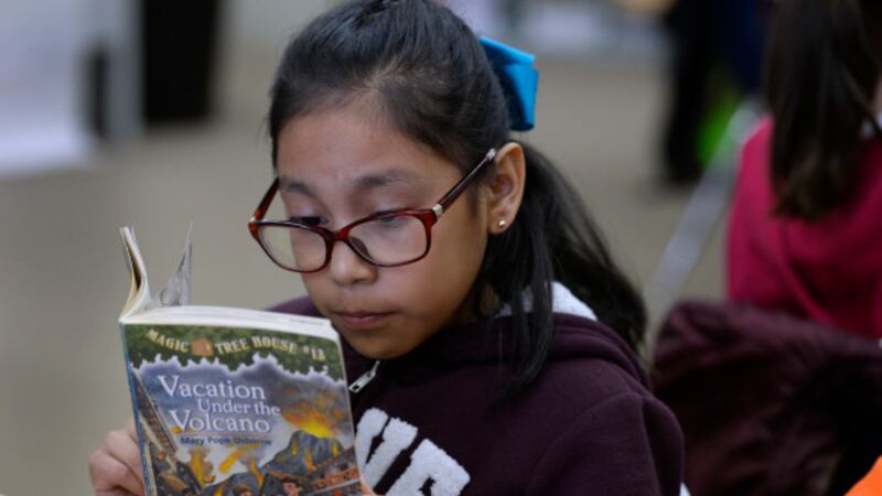 A 10-year-old girl wearing glasses reads “Vacation Under the Volcano” at a desk.