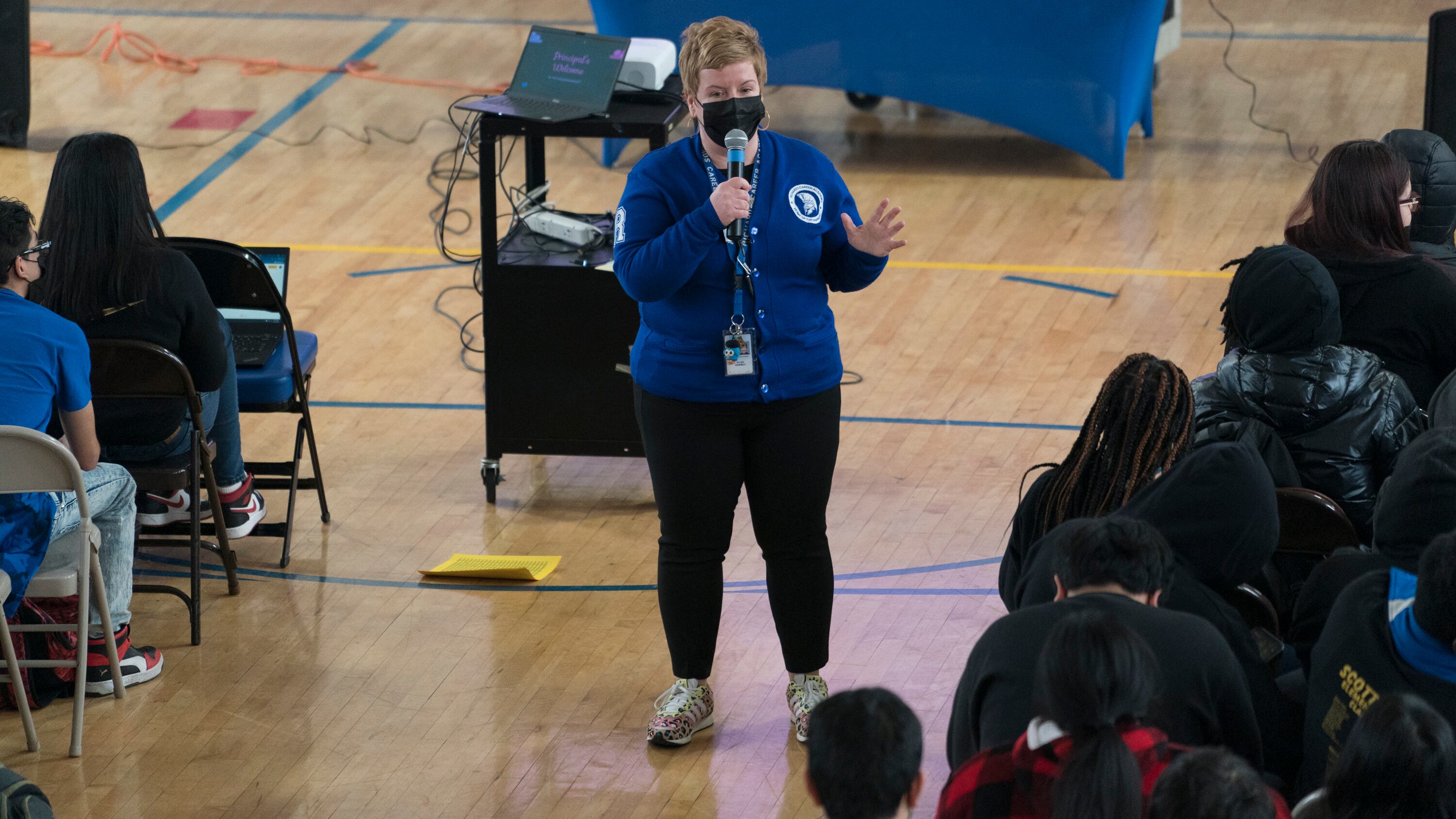 A school principal wearing a blue top and black pants addresses her students during an assembly in a high school gymnasium.
