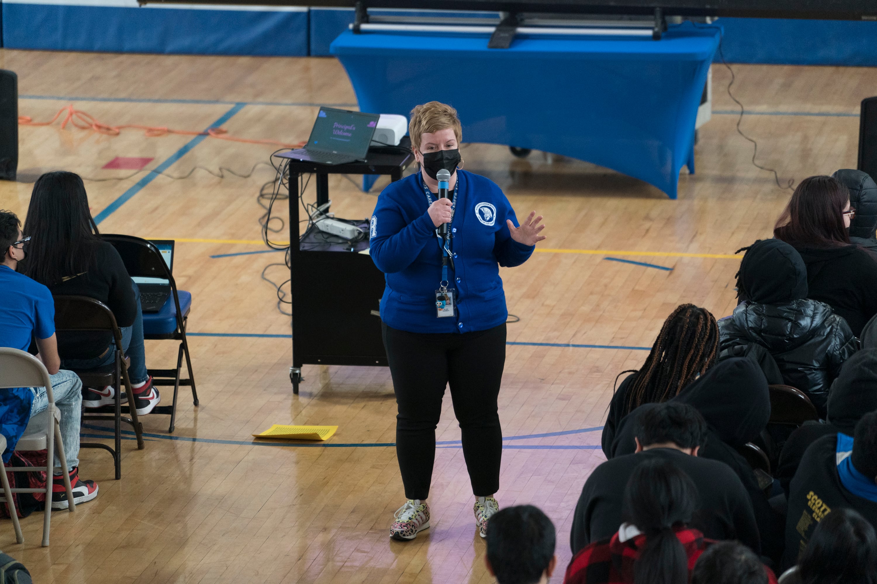 A school principal wearing a blue top and black pants addresses her students during an assembly in a high school gymnasium.