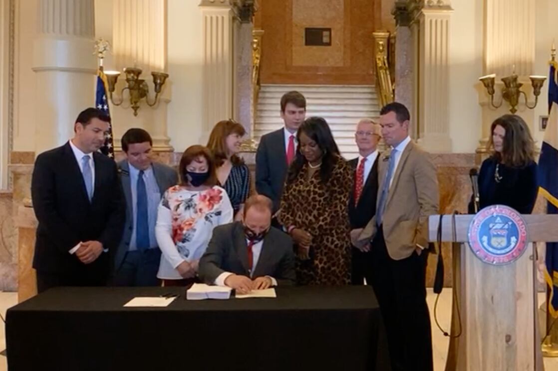 Seated at a table covered by a dark cloth, Gov. Jared Polis signs a piece of paper next to a large stack of documents. Colorado legislators stand around him looking on.