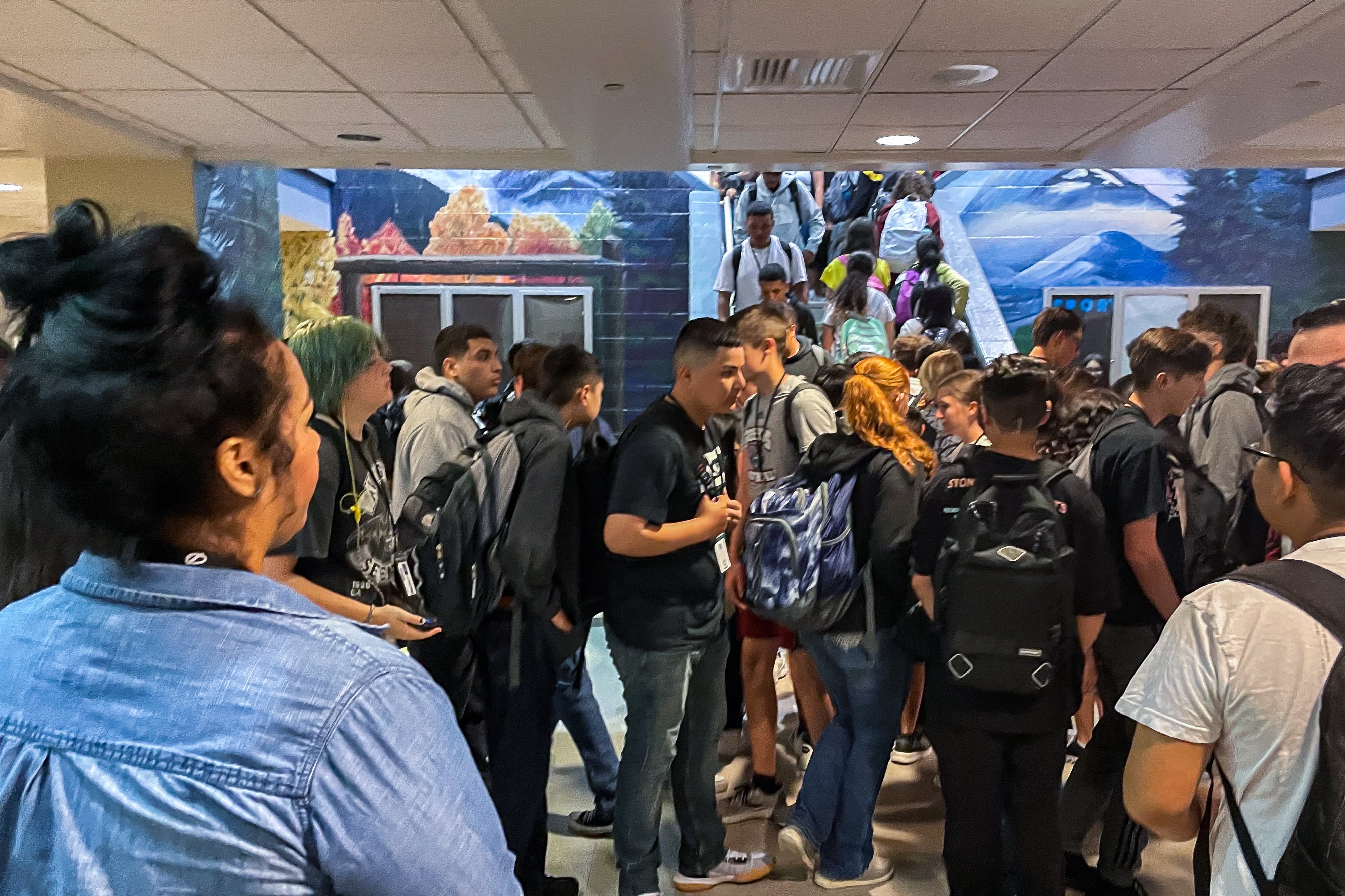 A woman with her back to the camera watches students in a crowded high school hallway. 