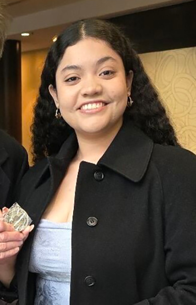 A high school senior poses for a portrait with long, dark, wavy hair and wearing a black jacket.