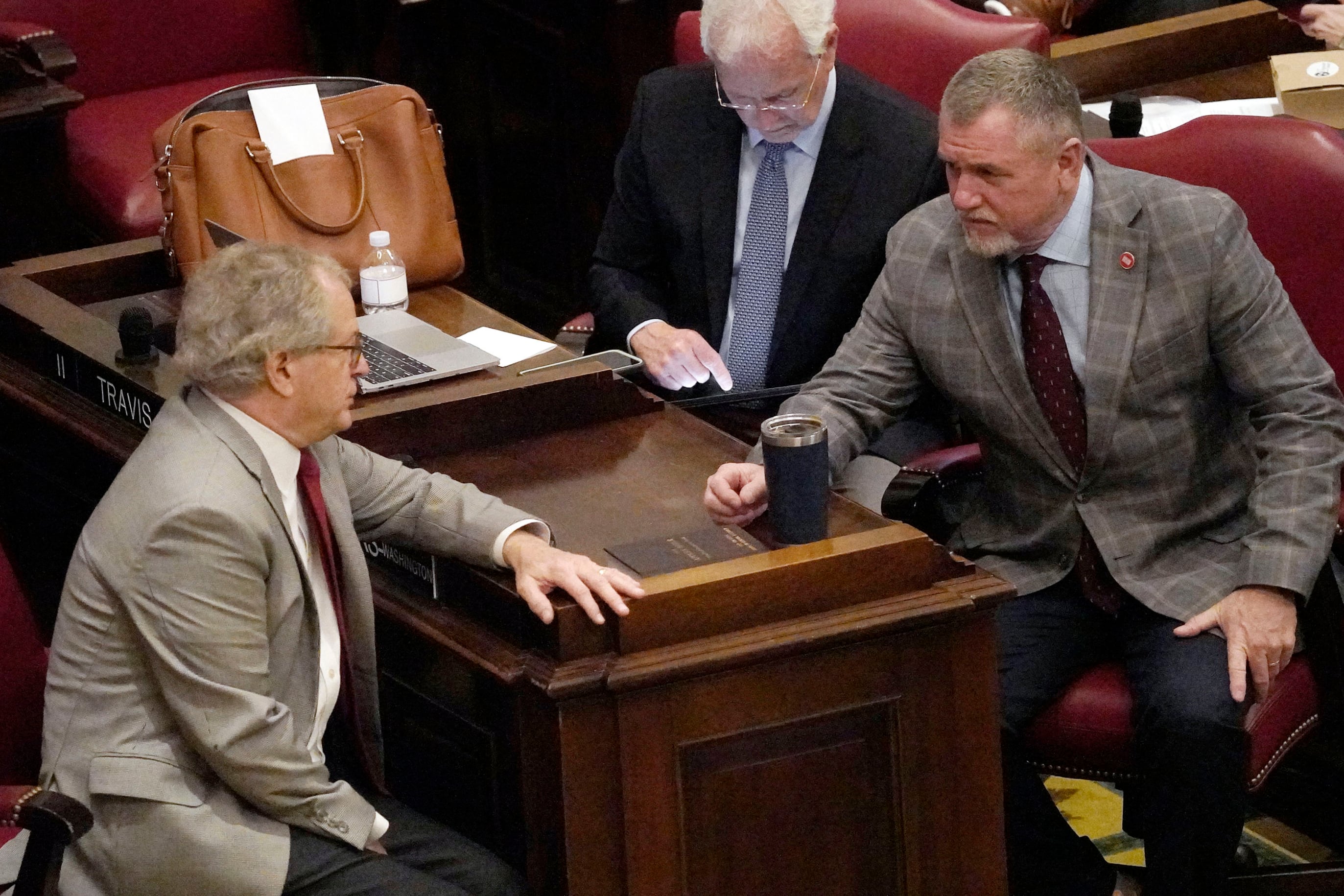 Several men wearing business suits talk around a desk.