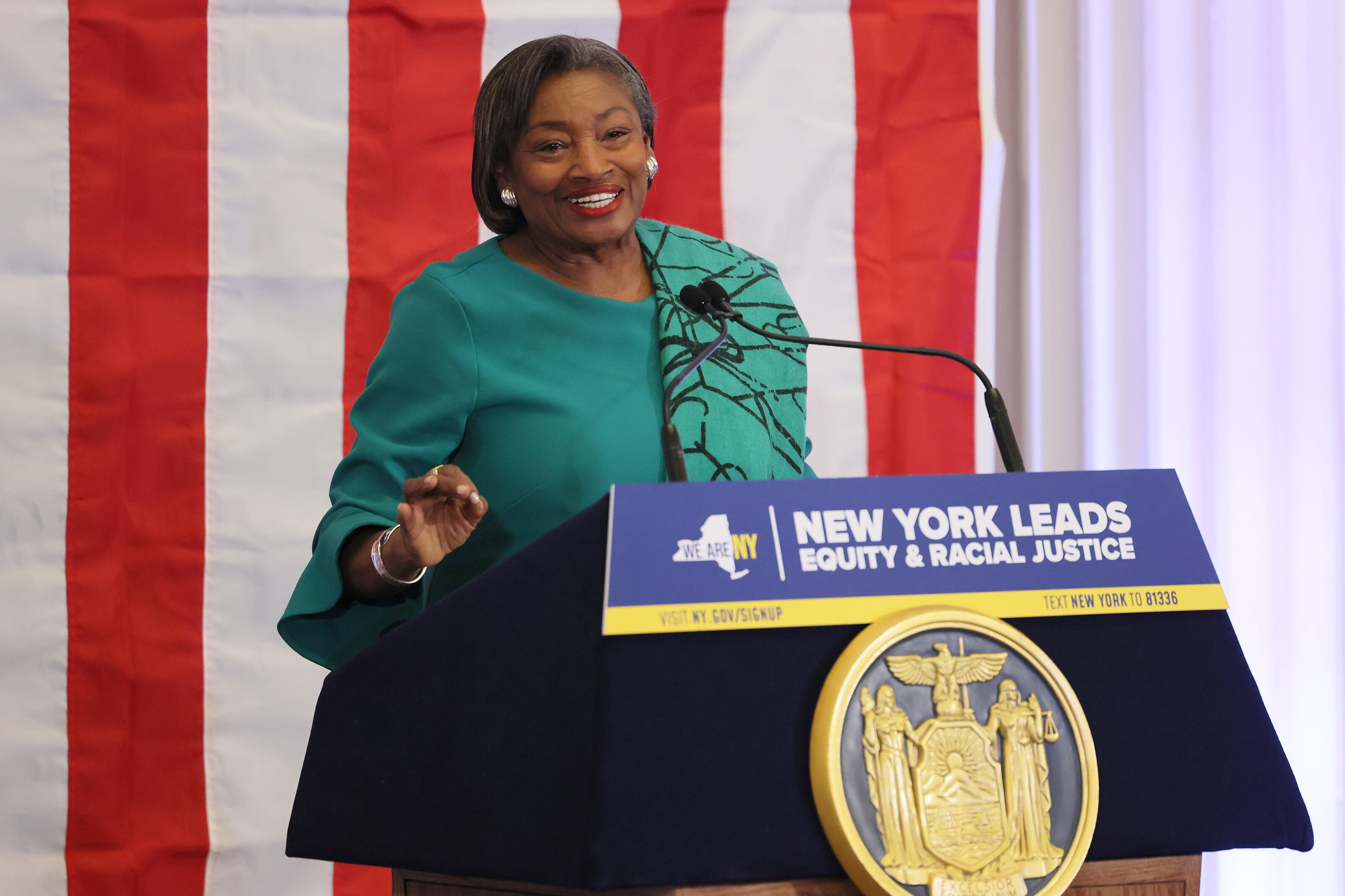A Black woman with short dark hair and wearing a green blouse speaks from behind a podium and in front of an American flag.