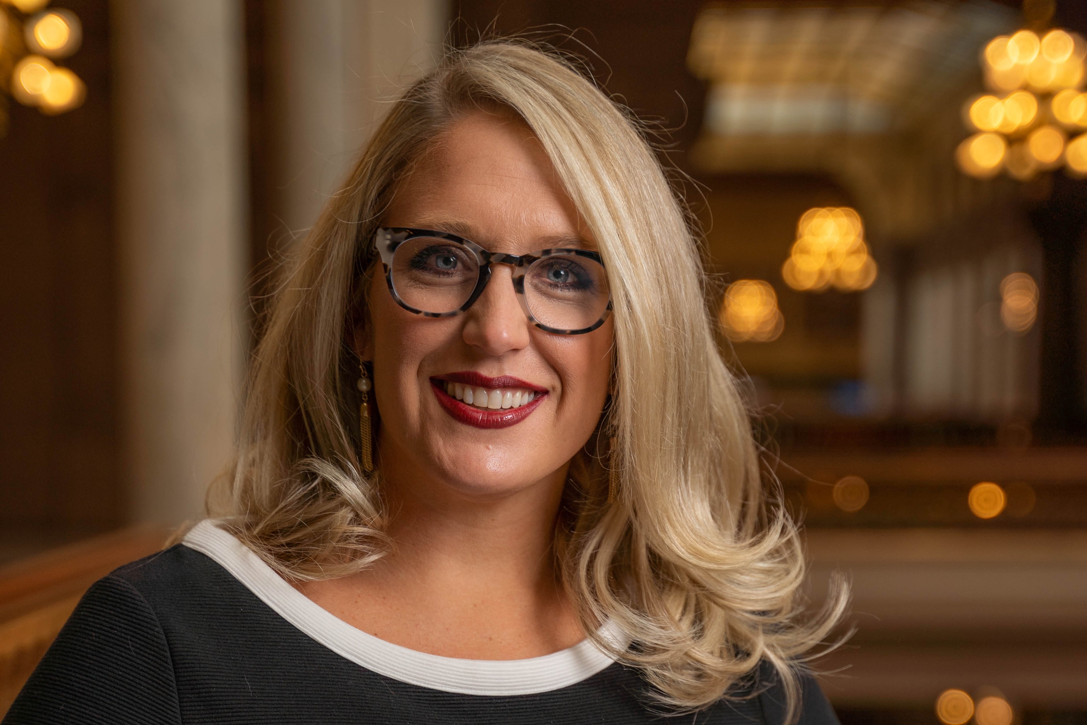 A woman with blonde hair and dark glasses wearing a dark shirt smiles at the camera.