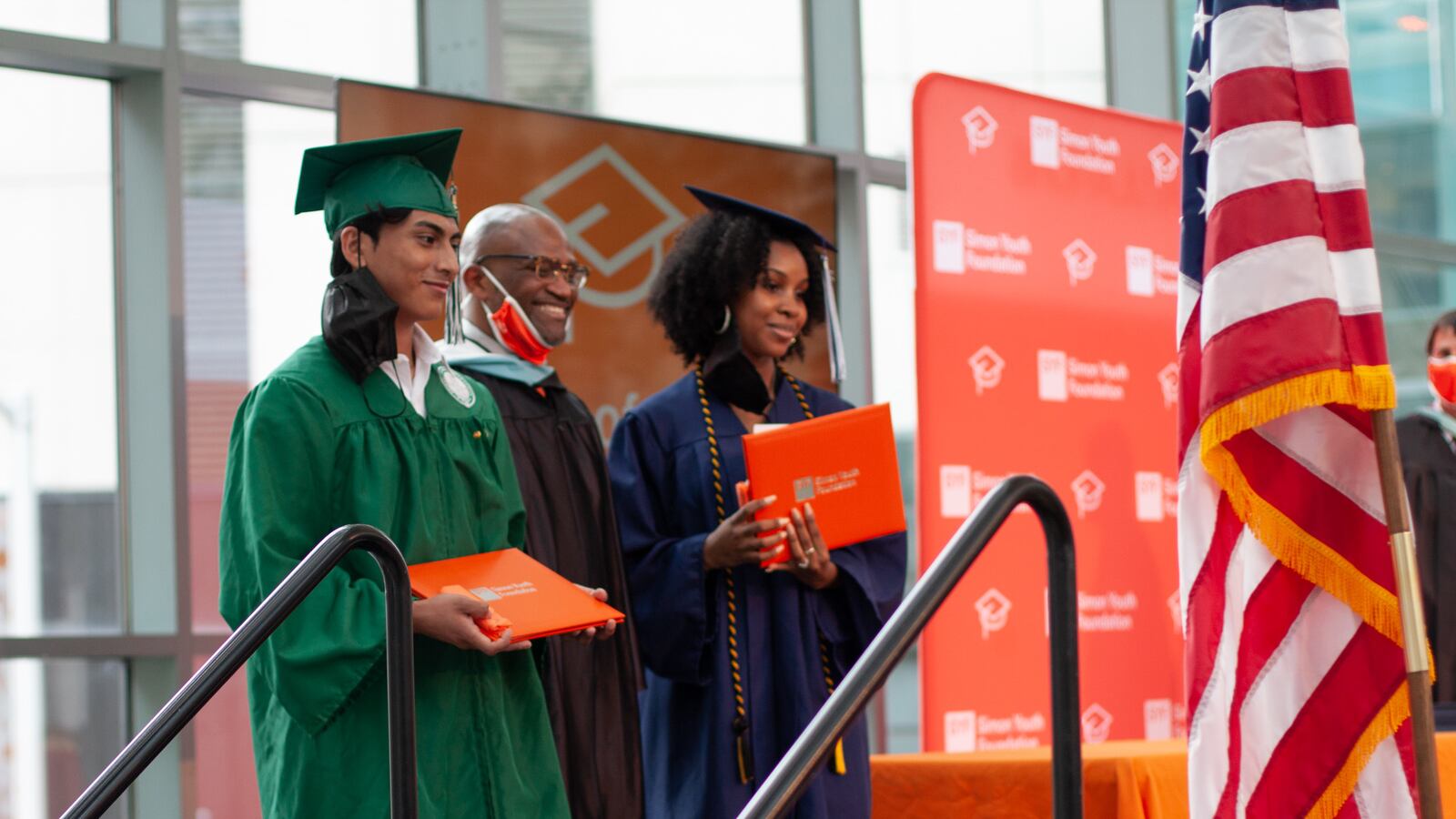 Two students, one wearing green graduation garb and the other wearing a blue cap and gown, hold orange award binders next to a their principal.