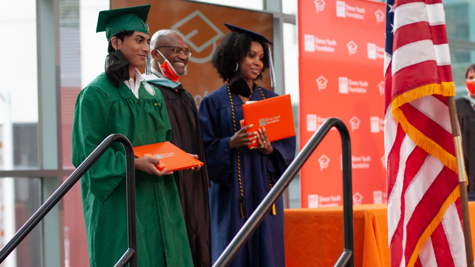 Two students, one wearing green graduation garb and the other wearing a blue cap and gown, hold orange award binders next to a their principal.