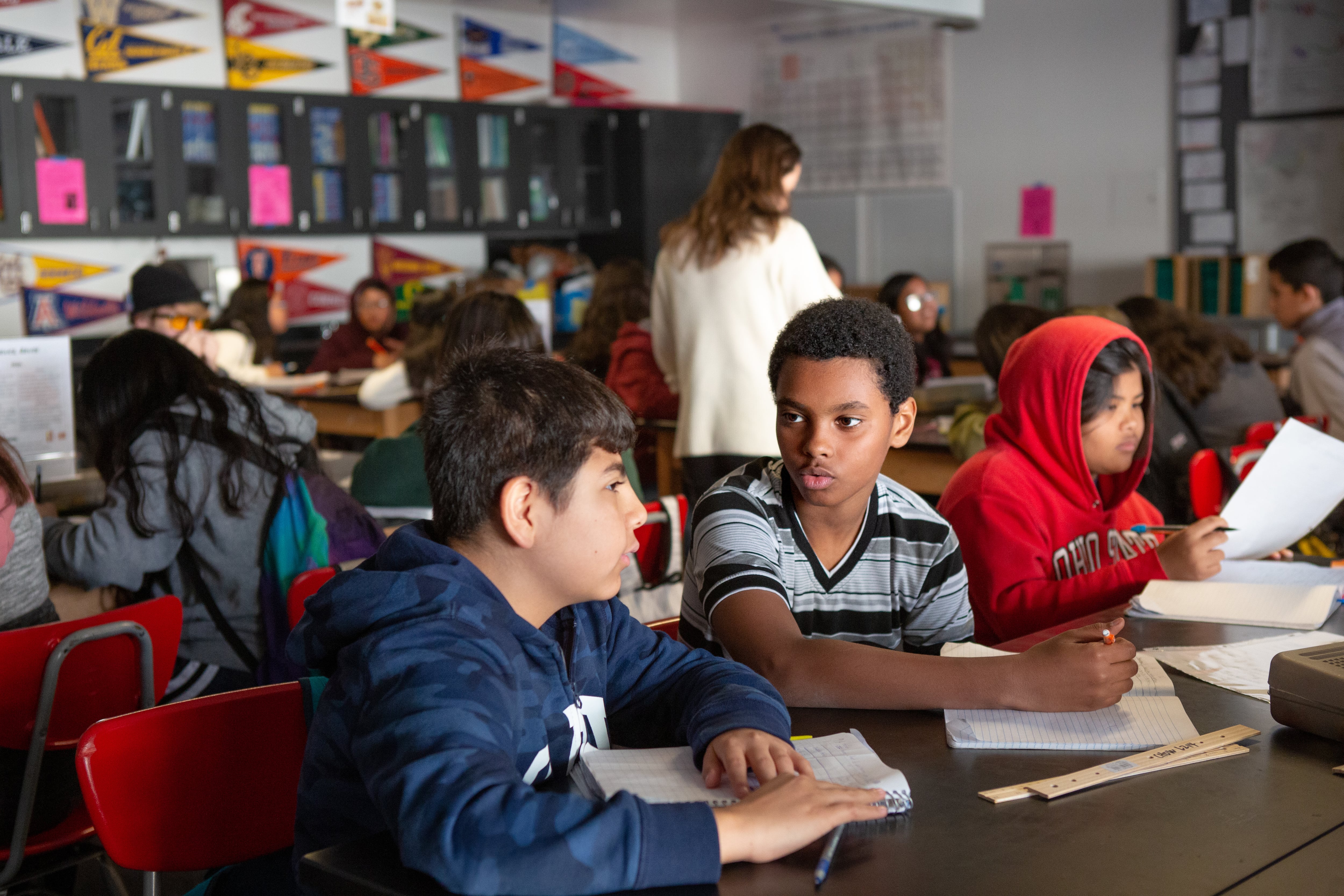 Students sit at a table with papers. A woman stands in the background.