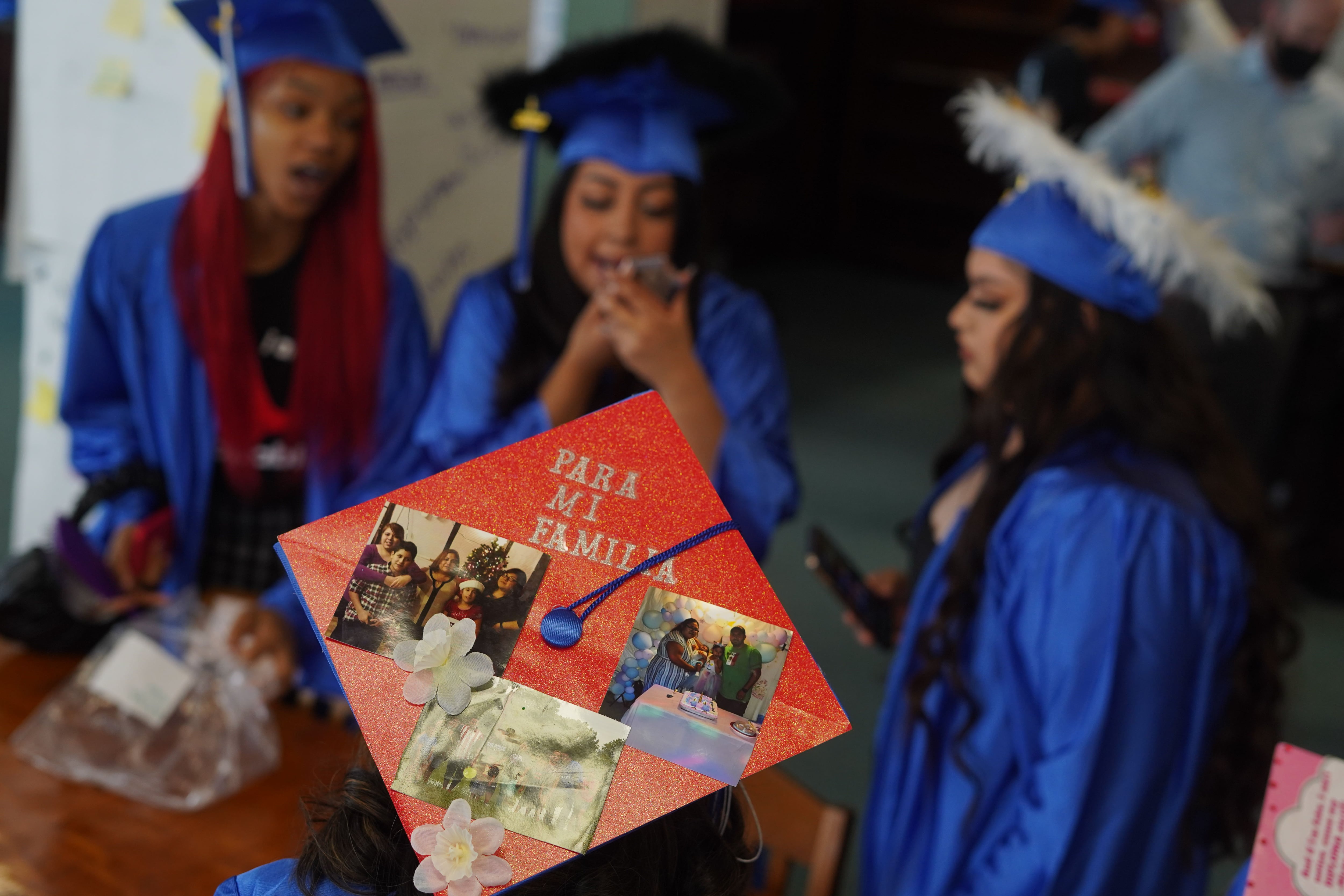 Three high school graduates in blue caps and gowns are in the background, while a close up of a graduation cap with photos taped to it is in the foreground.