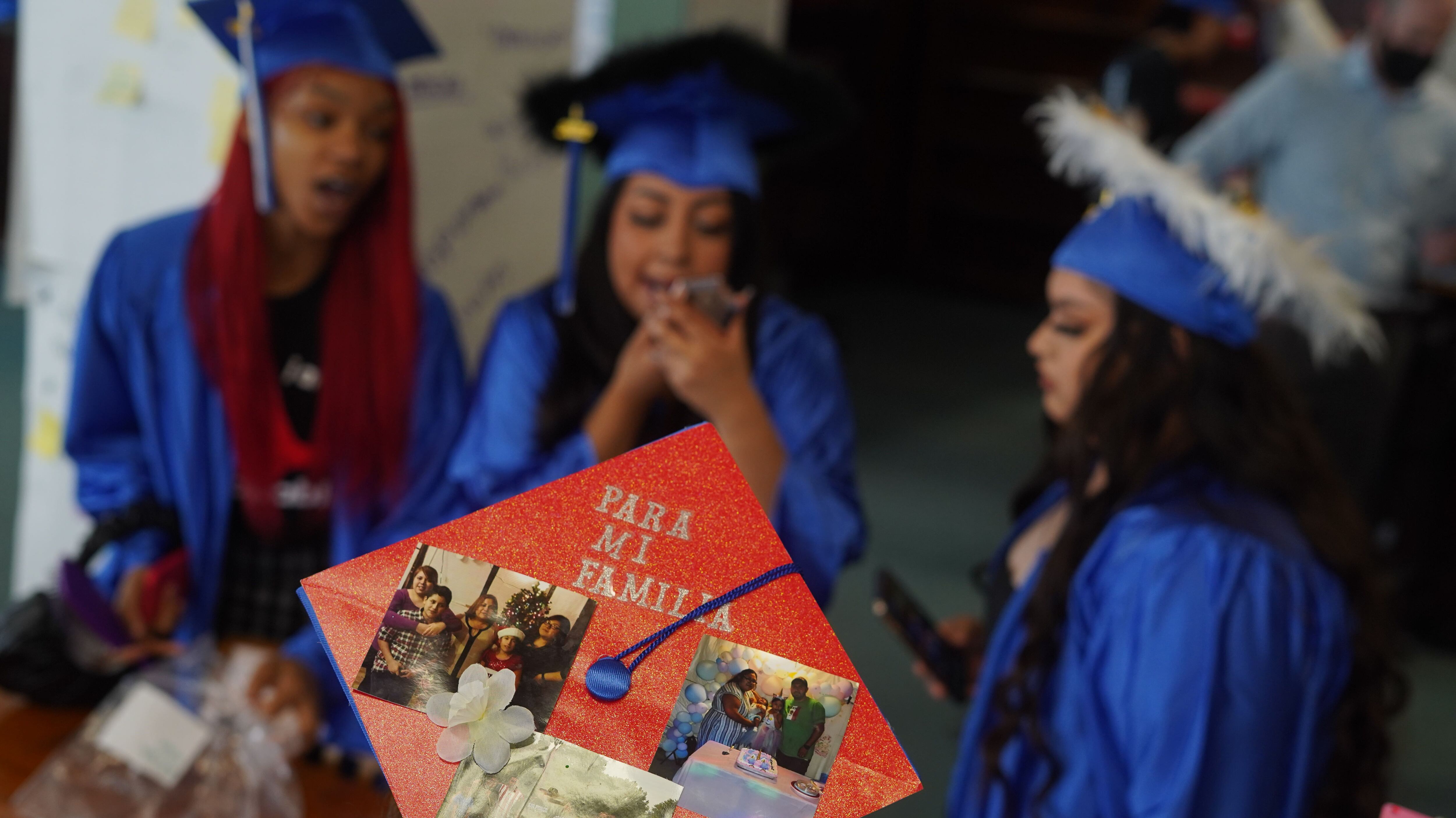 Three high school graduates in blue caps and gowns are in the background, while a close up of a graduation cap with photos taped to it is in the foreground.