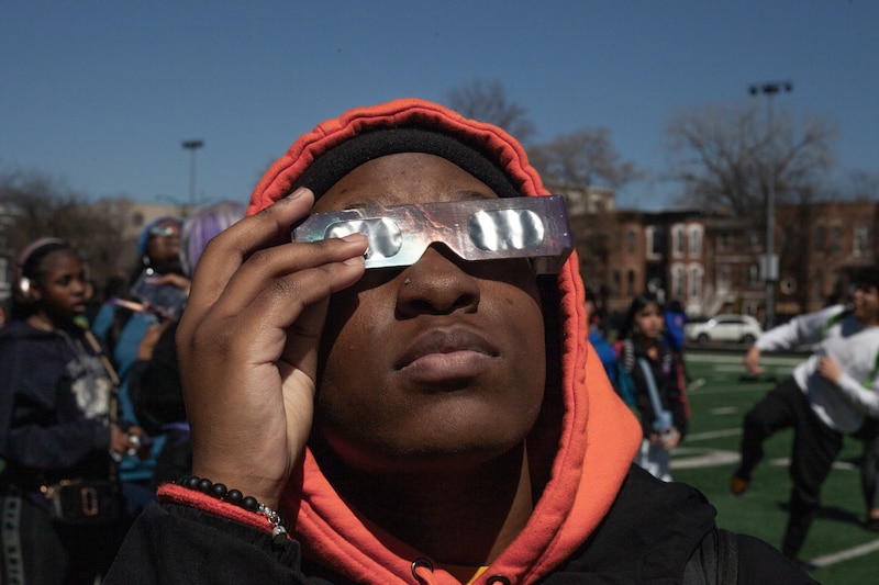 A student wearing a coloful hoodie and holding a pair of eclipse viewing glasses looks up at the sky with other students in the background.