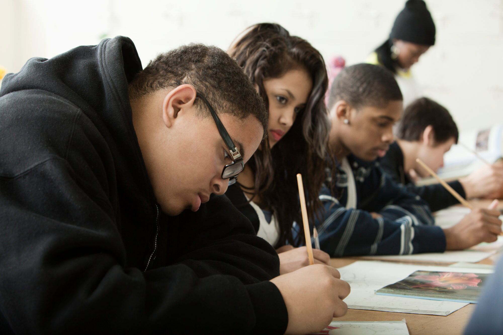 Four students take a written test on a wooden desk with a teacher standing in the background.
