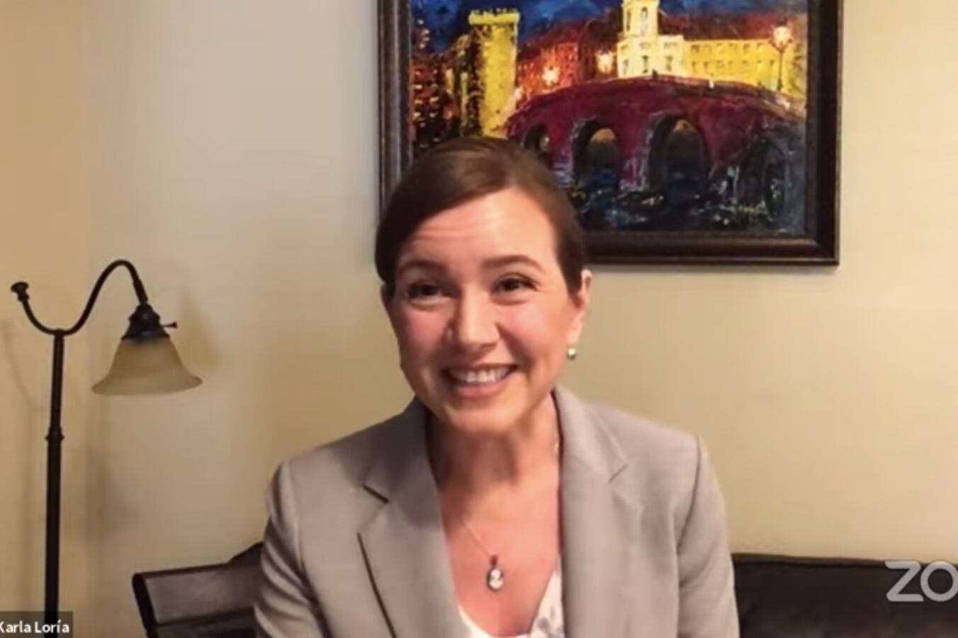 Karla Loria, a woman in a beige suit, sits, smiling, in the center with a painting on the wall behind her.
