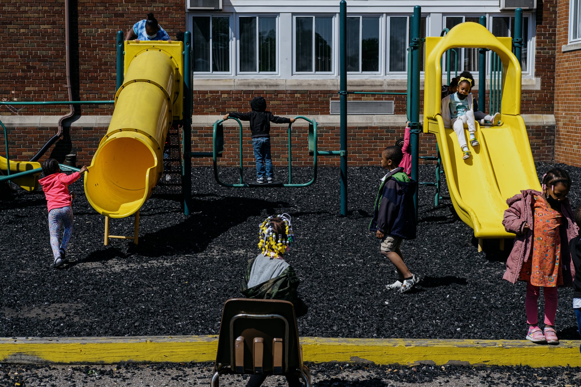 A group of young students play on a playground set outside with a brick school building in the background.