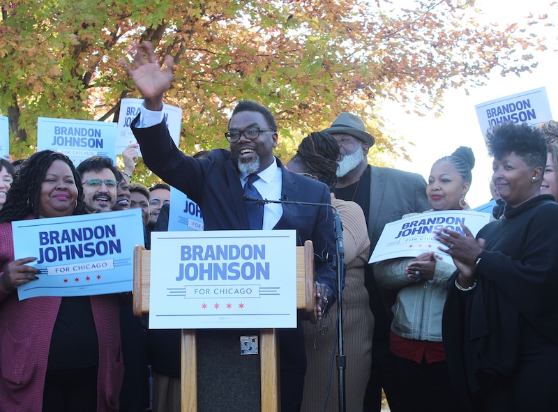 Man in suit raises right hand surrounded by supporters