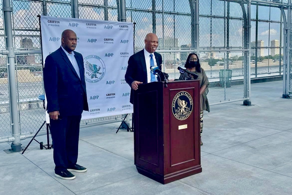 A man stands at a lectern outside, flanked by another man on his left and a woman on his right.