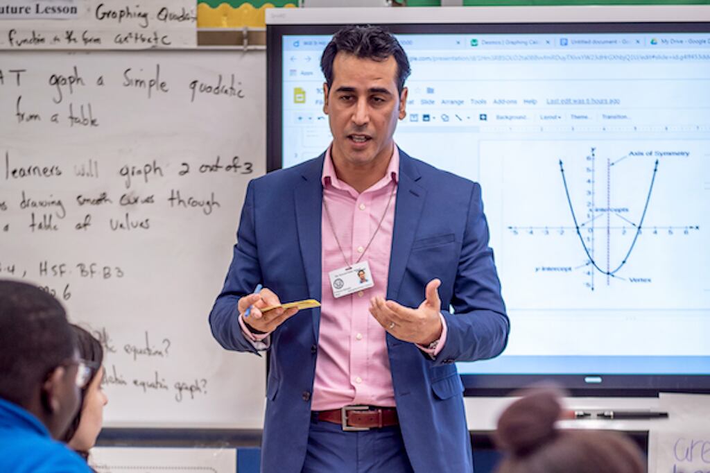 A male teacher in a blue suit with a pink collared shirt stands at the front of his class facing his students as a math problem is projected on the white board behind him.