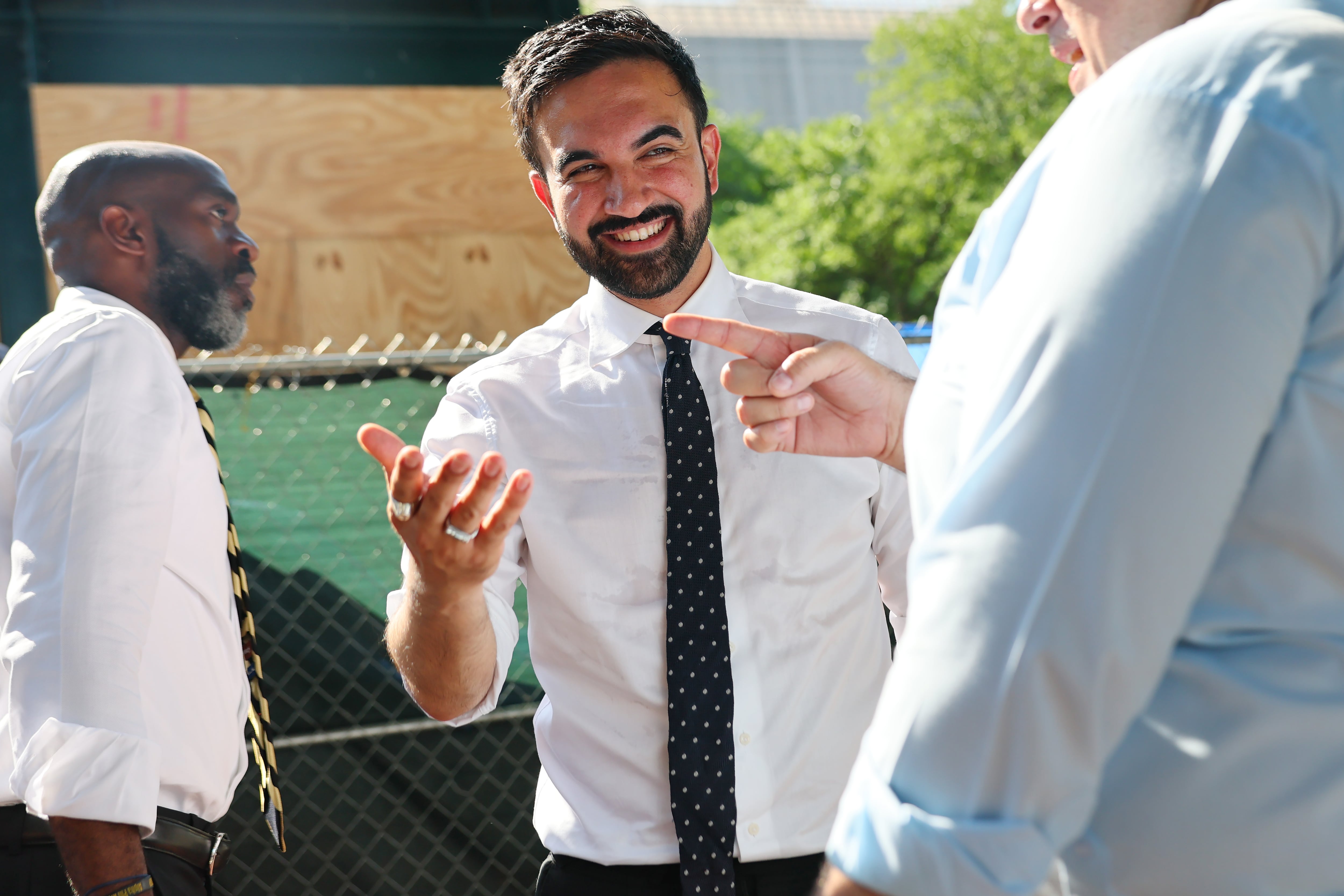 A man in a white shirt and dark tie smiles and holds his right hand up.