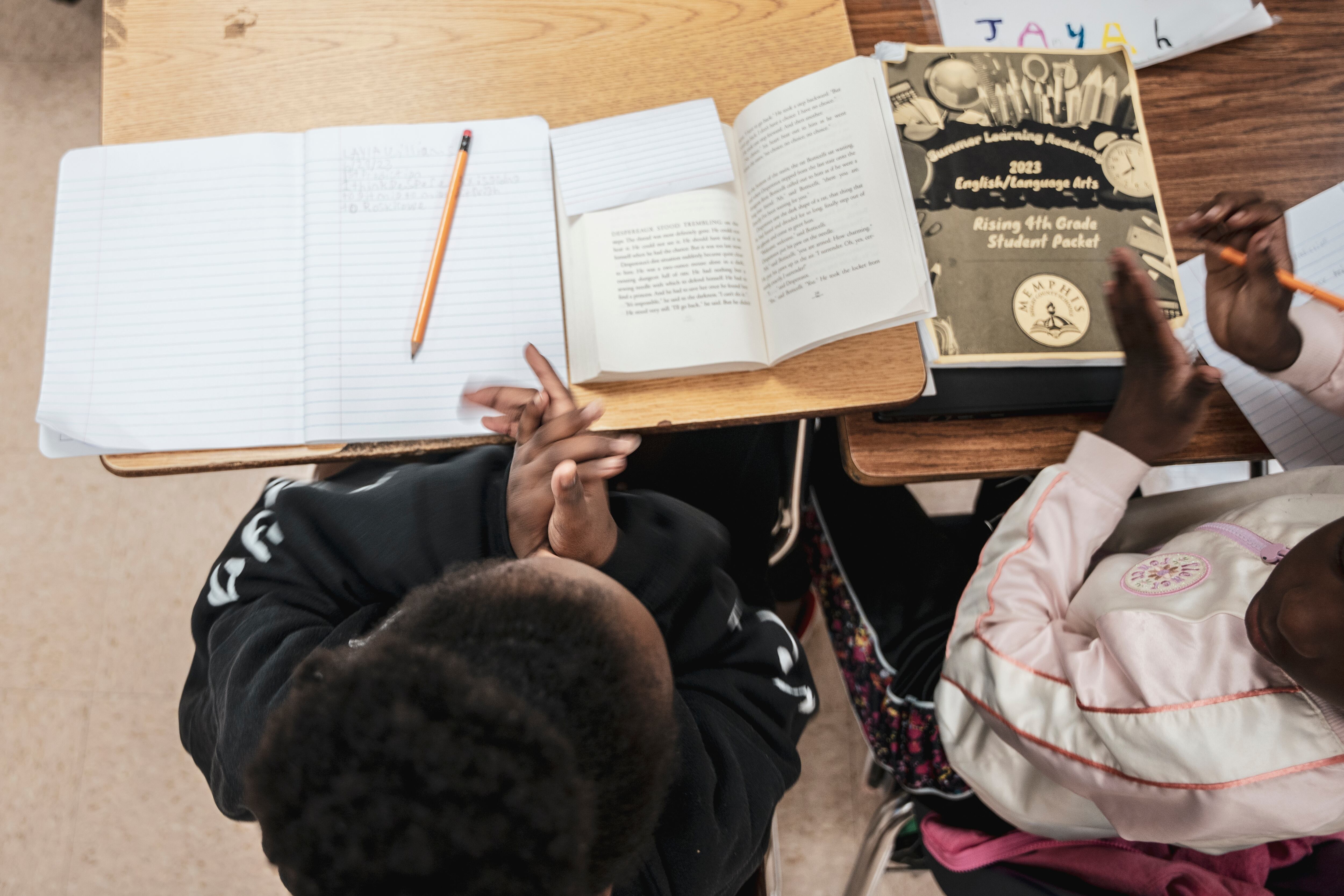 Two students clap while sitting at desks with notebooks, paper, and a pencil in front of them.