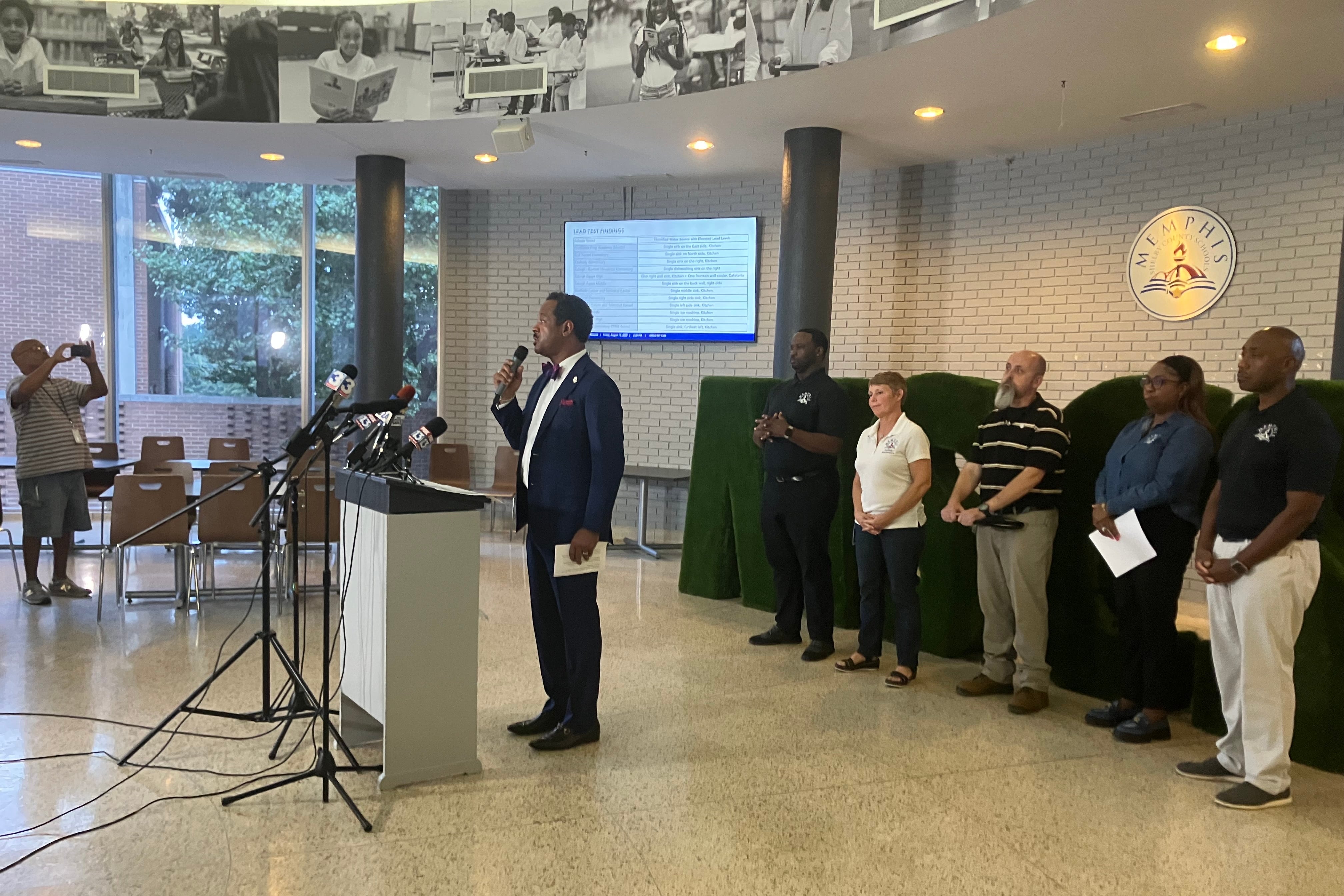 Superintendent Roderick Richmond stands with a microphone in front of a podium, talking. Behind him are various district officials standing in a line.