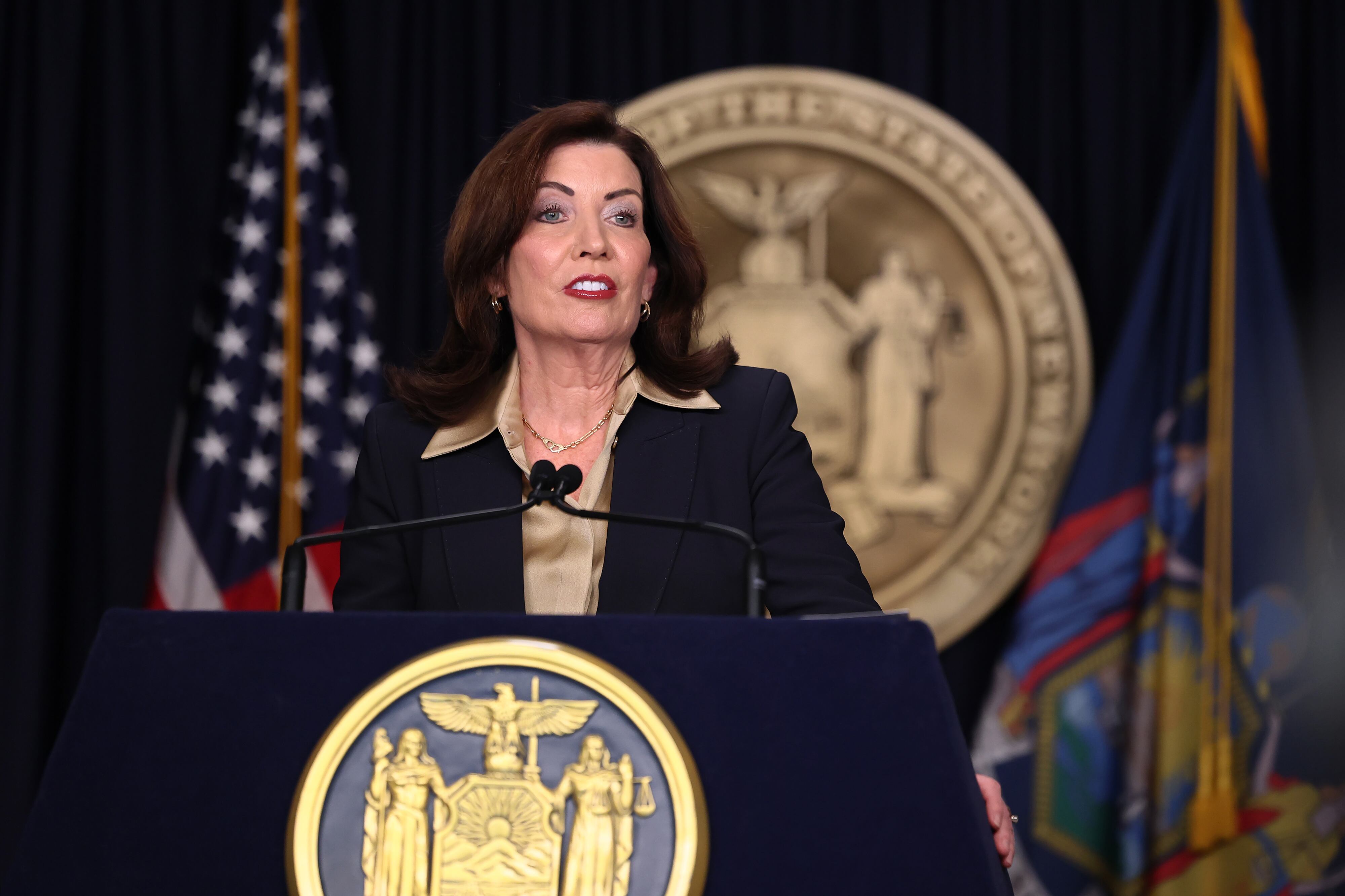 A white woman with dark hair and wearing a suit stands in front of a NY state seal and two flags and speaks from a podium.