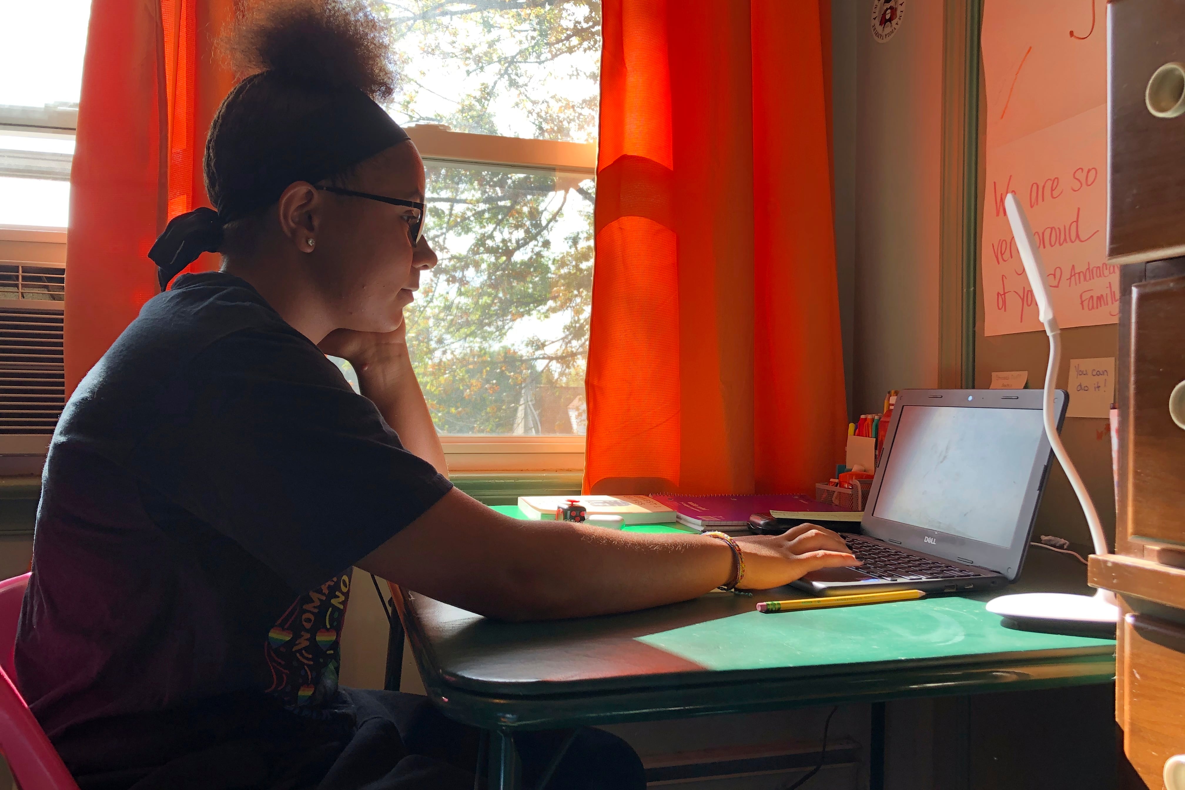 A teenage girl participates in a remote class on her laptop, sitting at a desk in her room.