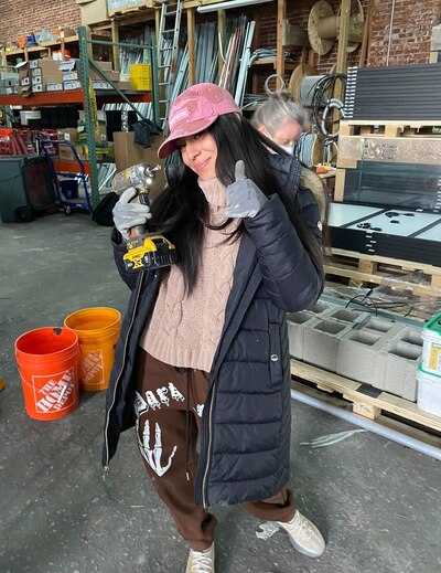 A young woman with long dark hair and wearing a black puffy jacket and a pink hat poses for a photo holding a drill.