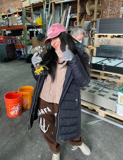 A young woman with long dark hair and wearing a black puffy jacket and a pink hat poses for a photo holding a drill.
