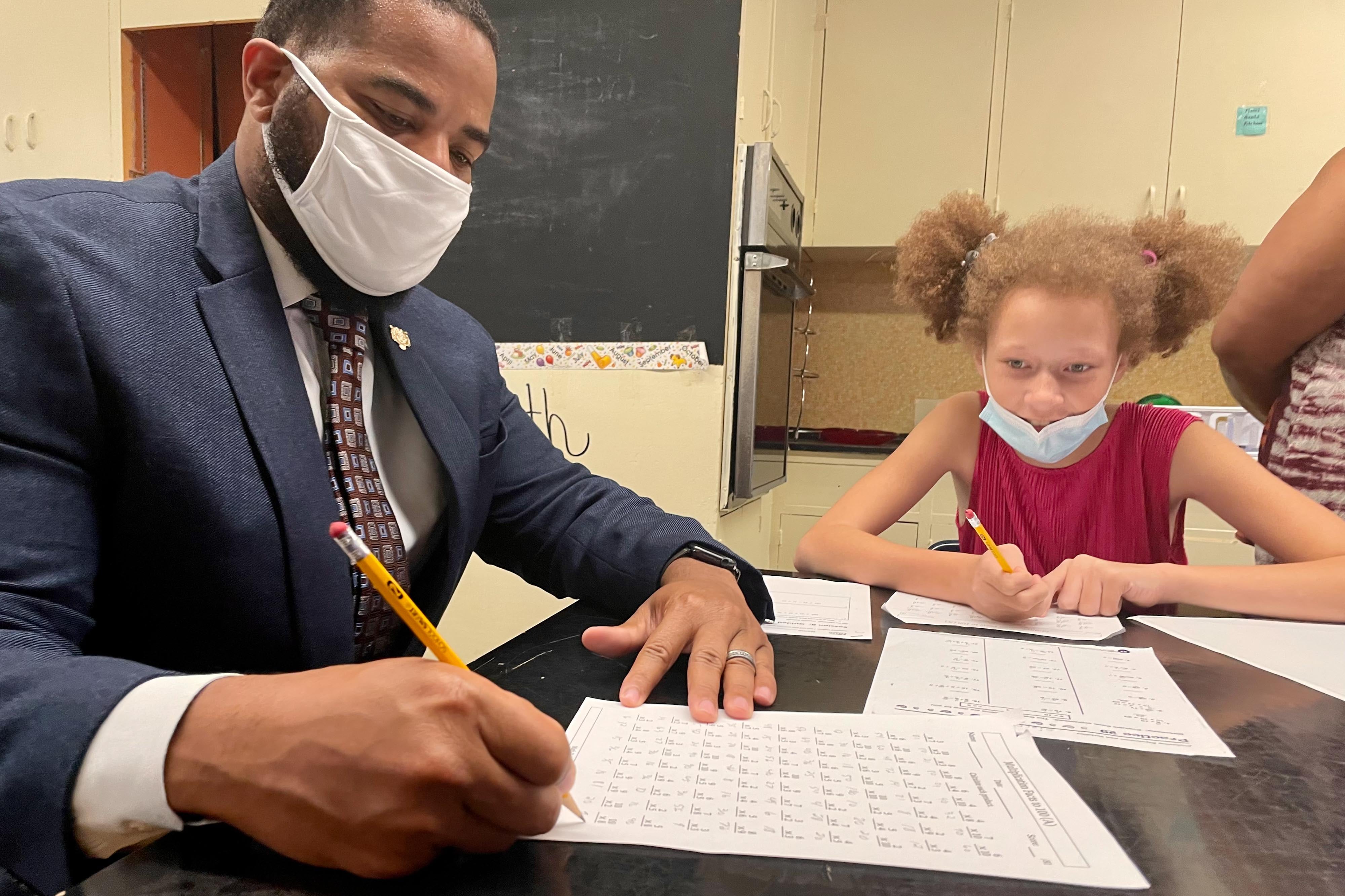 A male school administrator in a suit helps a girl with a multiplication worksheet, sitting with her at a desk. They are both wearing masks to prevent the spread of COVID.