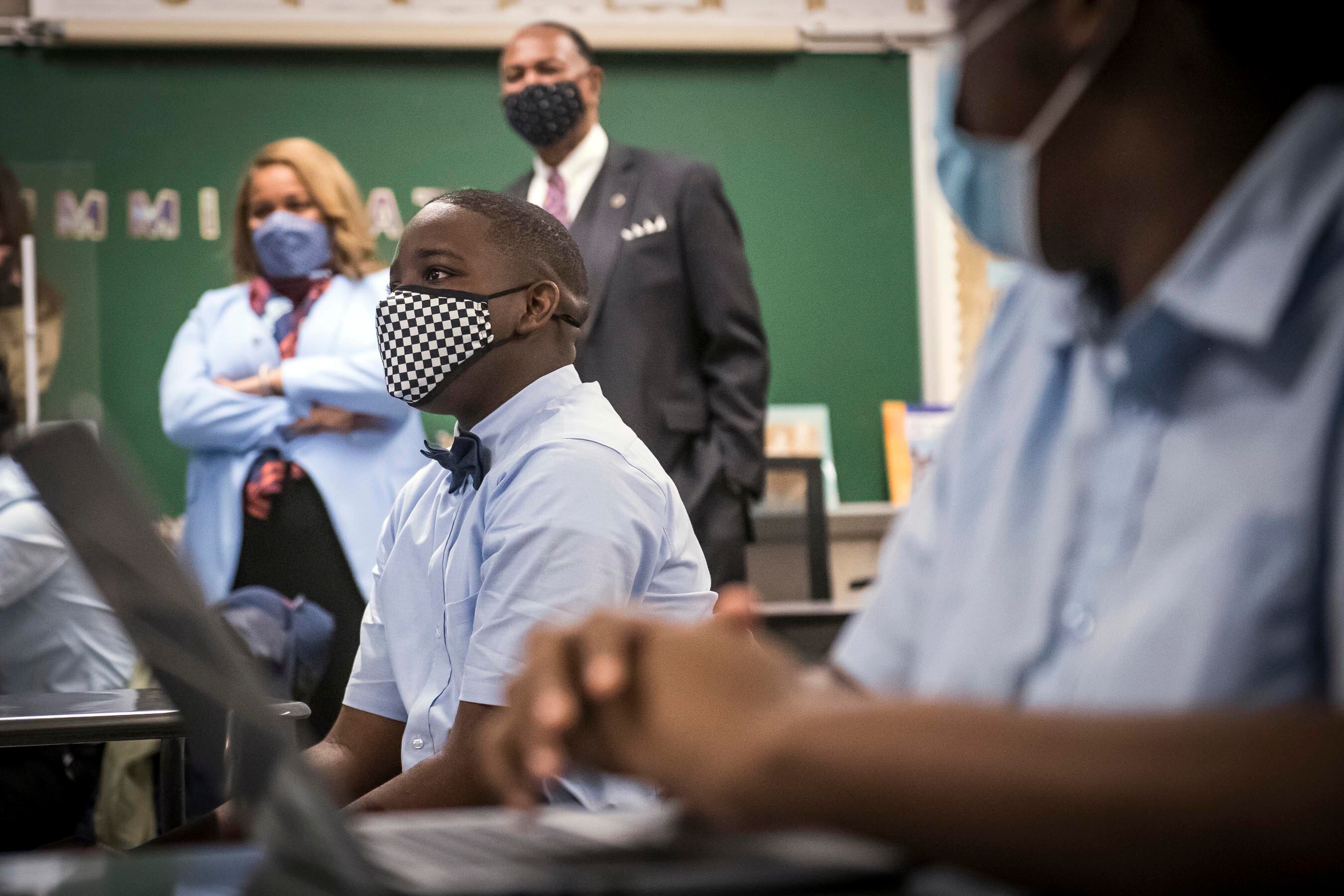 Students in blue shirts and protective masks look toward the front of the classroom as Commissioner Porter observes in the background.