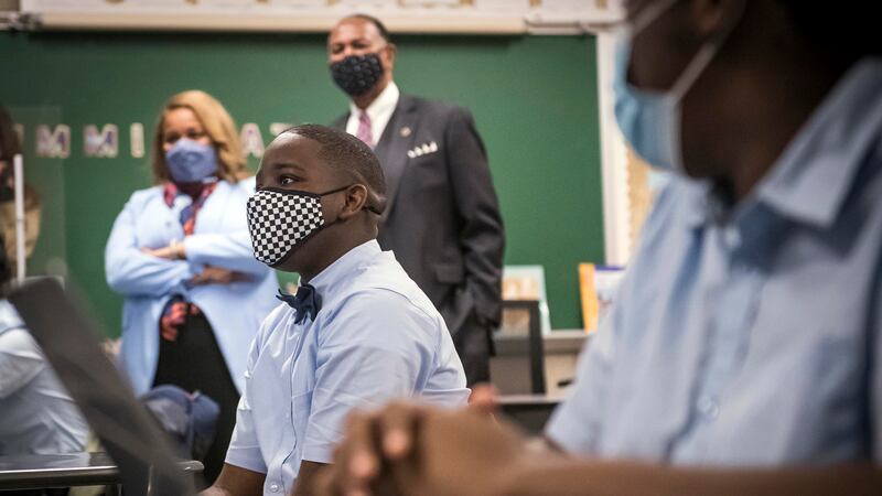 Students in blue shirts and protective masks look toward the front of the classroom as Commissioner Porter observes in the background.