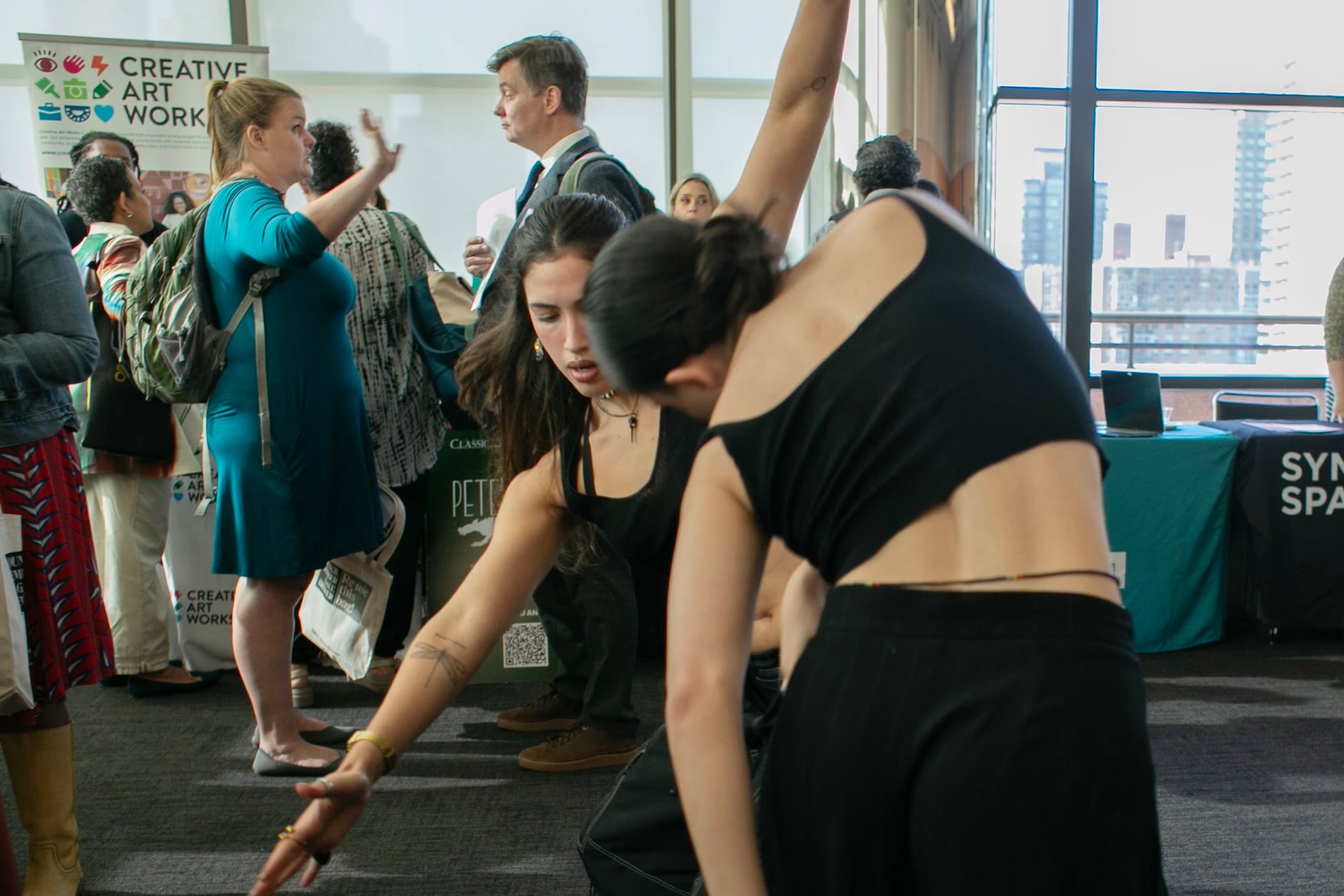 Two young women in black leotards dance in a room filled with people.