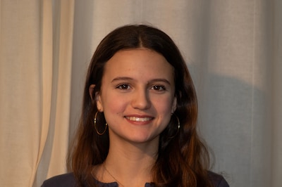 A photograph of a high school junior with long dark hair and wearing earrings poses for a portrait against a white cloth.