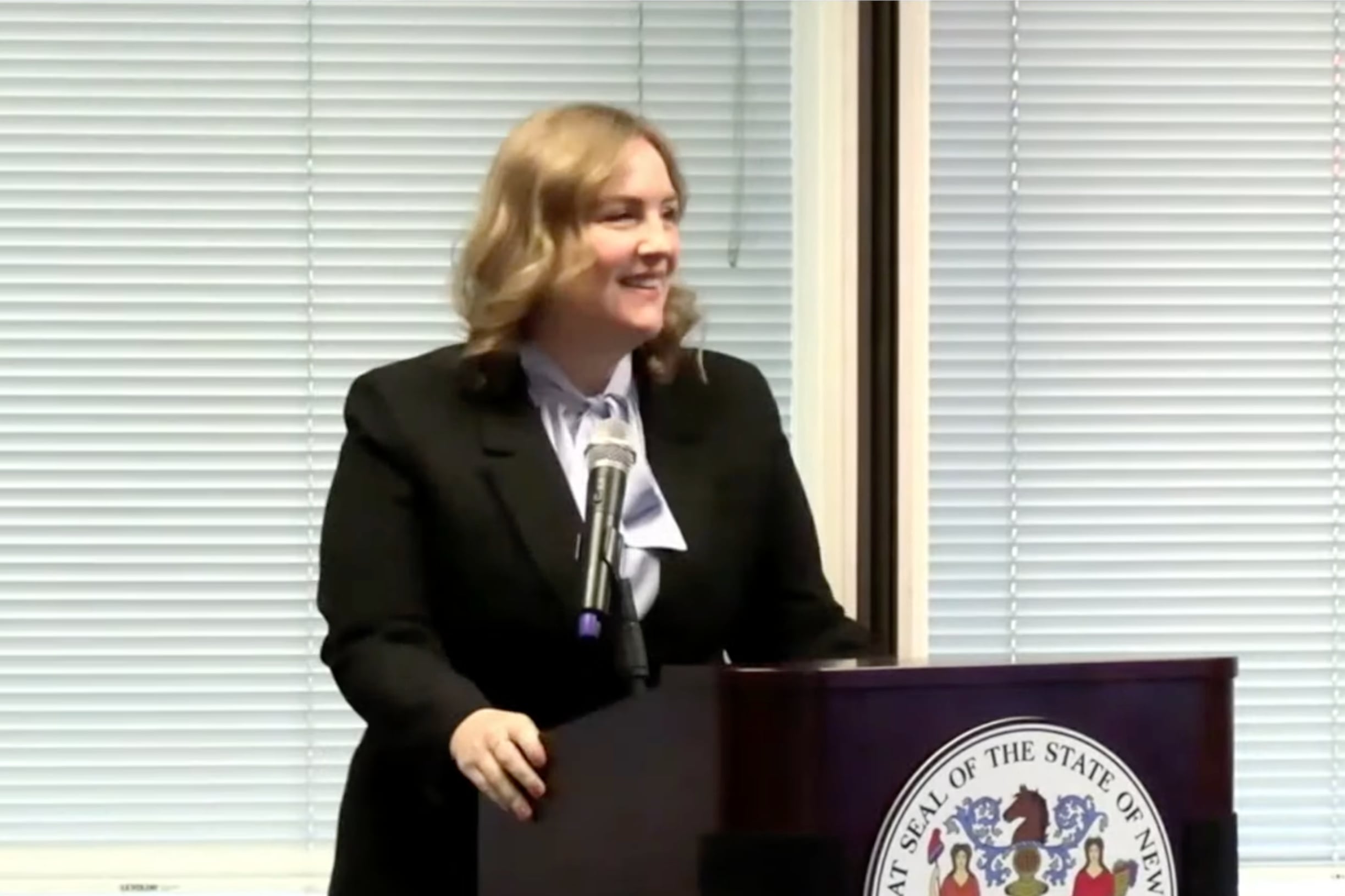 A woman with blonde hair stands behind a podium as she speaks into a microphone.