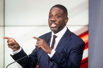 A photograph of a Black man in a dark blue suit speaking from behind a podium during a debate.