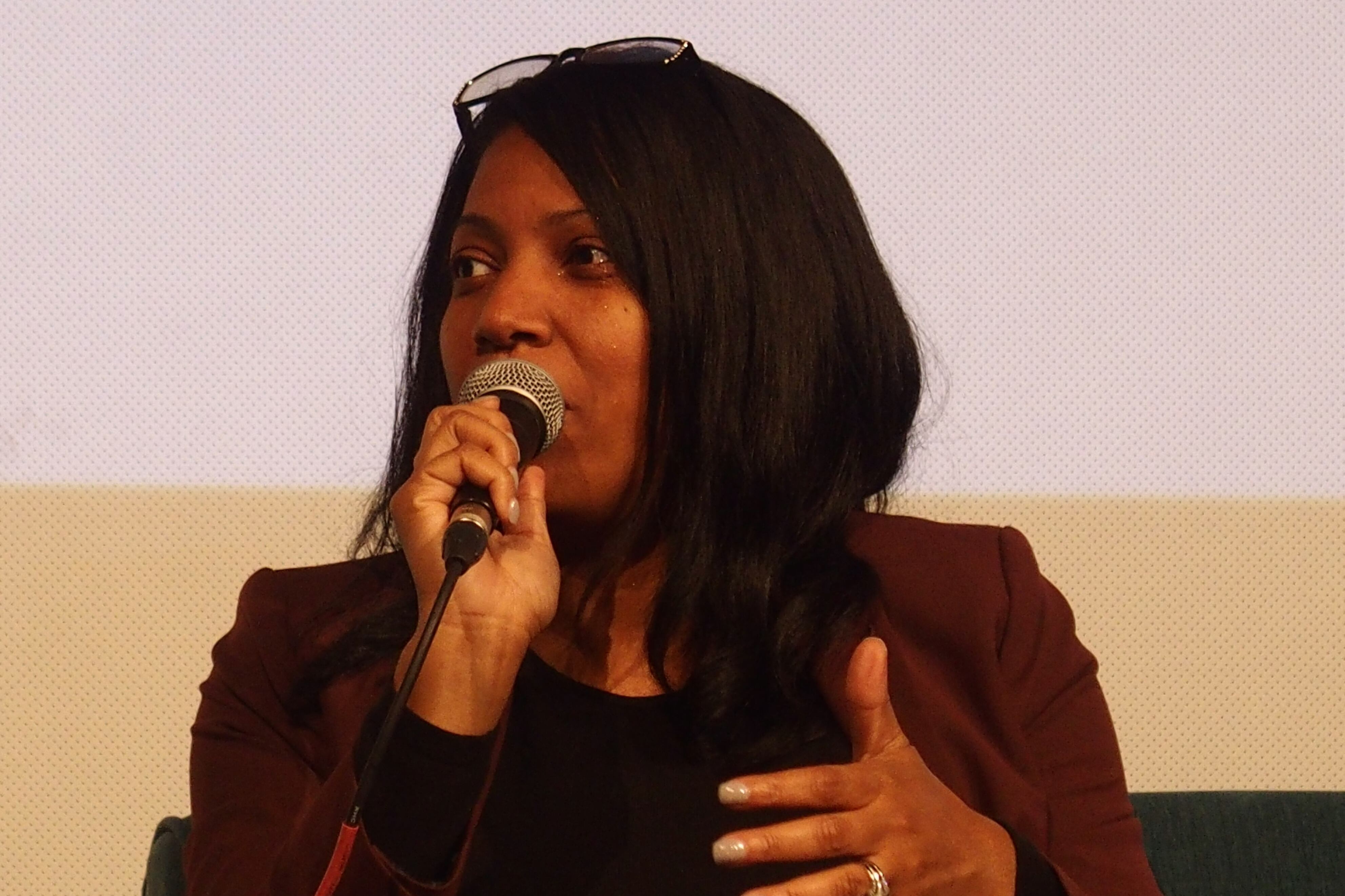 A woman with long dark hair and sunglasses on her head speaks into a microphone. She is wearing black clothes.