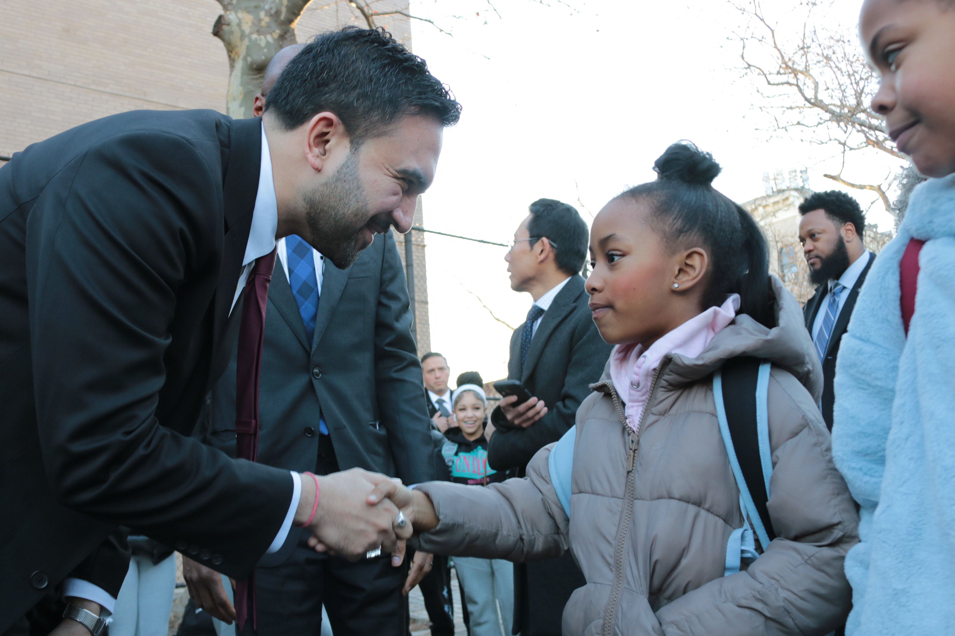 A photograph of a man in a suit shaking a hand of a young Black student wearing a jacket and a backpack with a large crowd of students in the background.