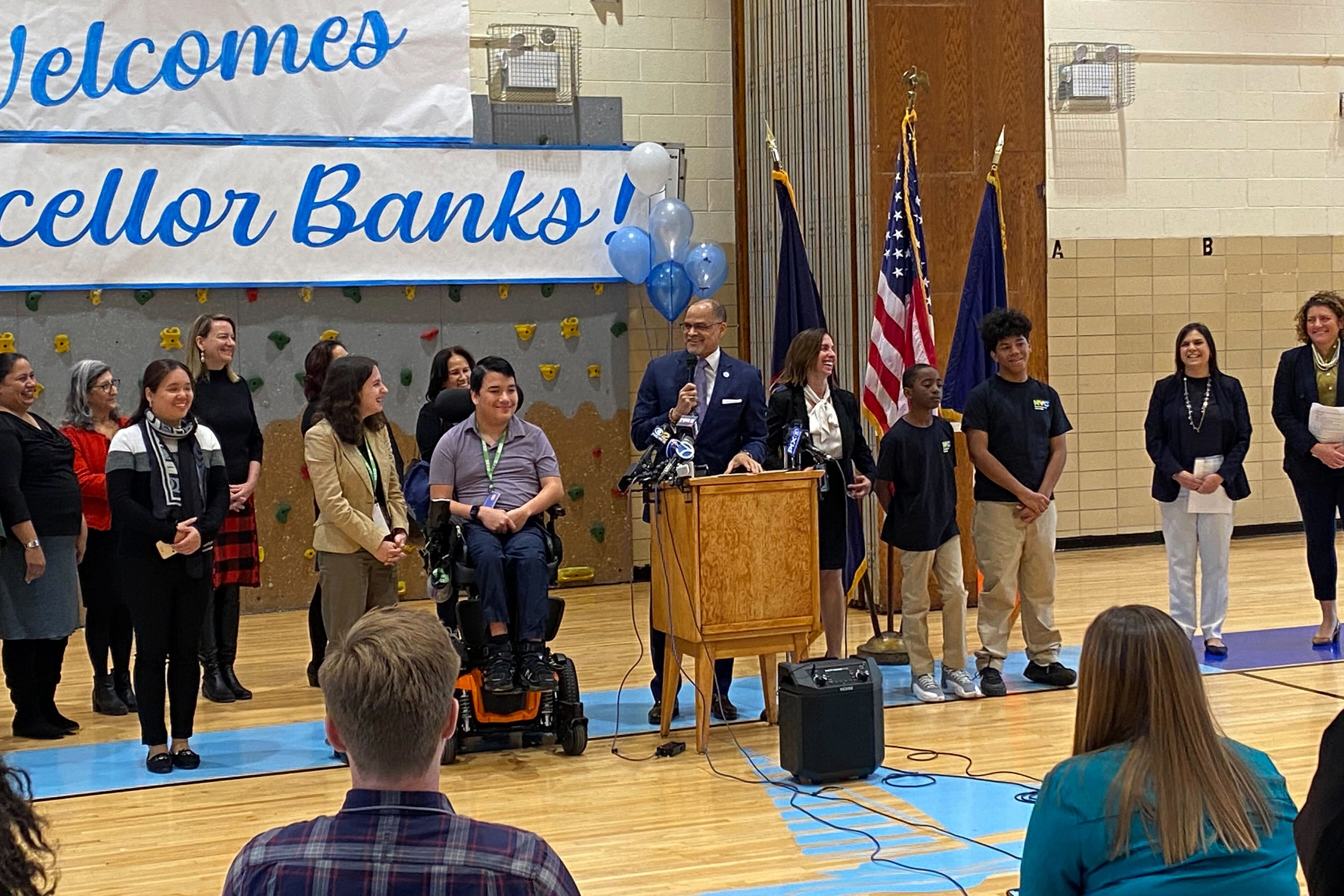 A group of people stand in front of a crowd in a school gym with flags and a banner in the background.