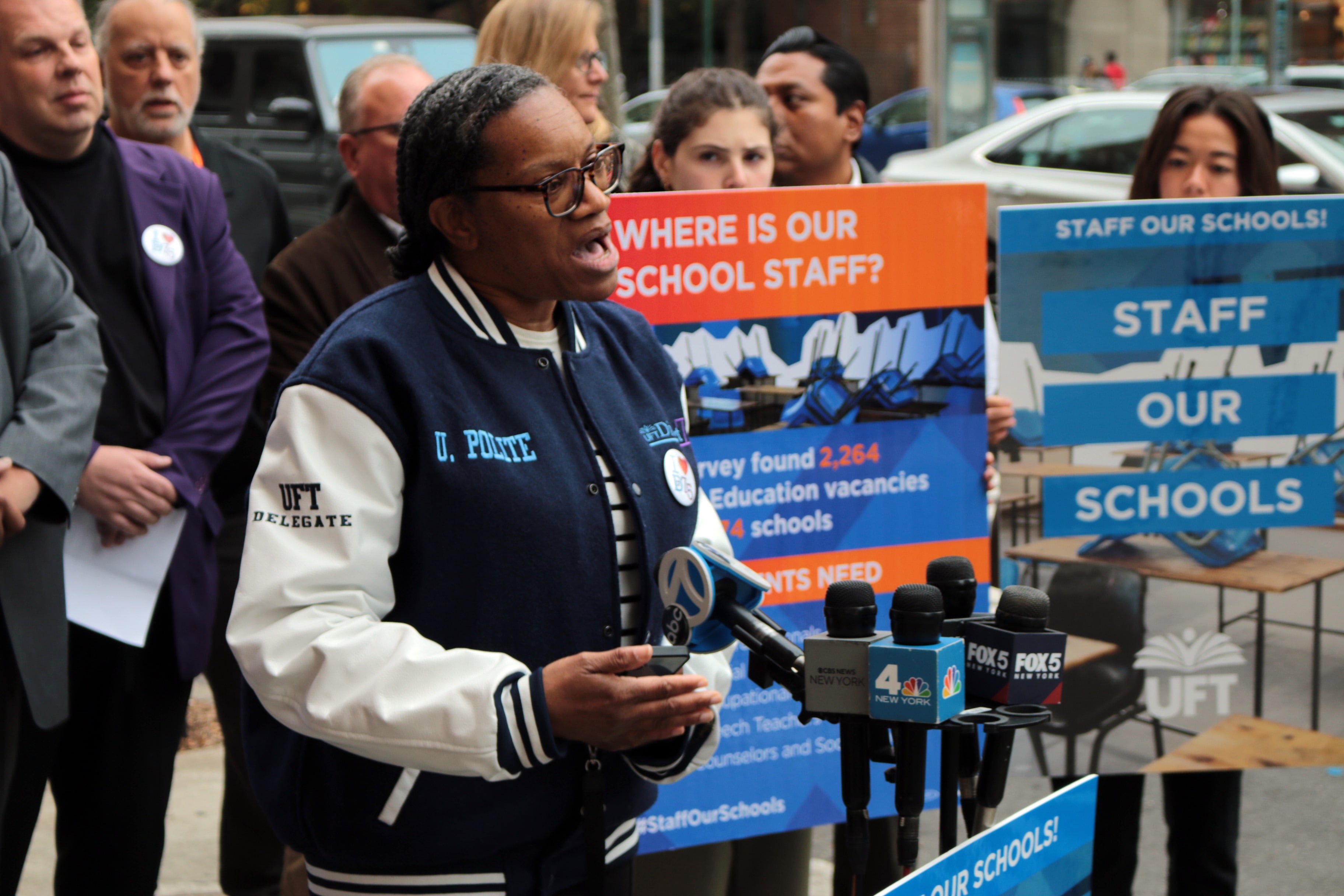 A person speaks from behind a row of microphones and in front of a group of people holding signs.