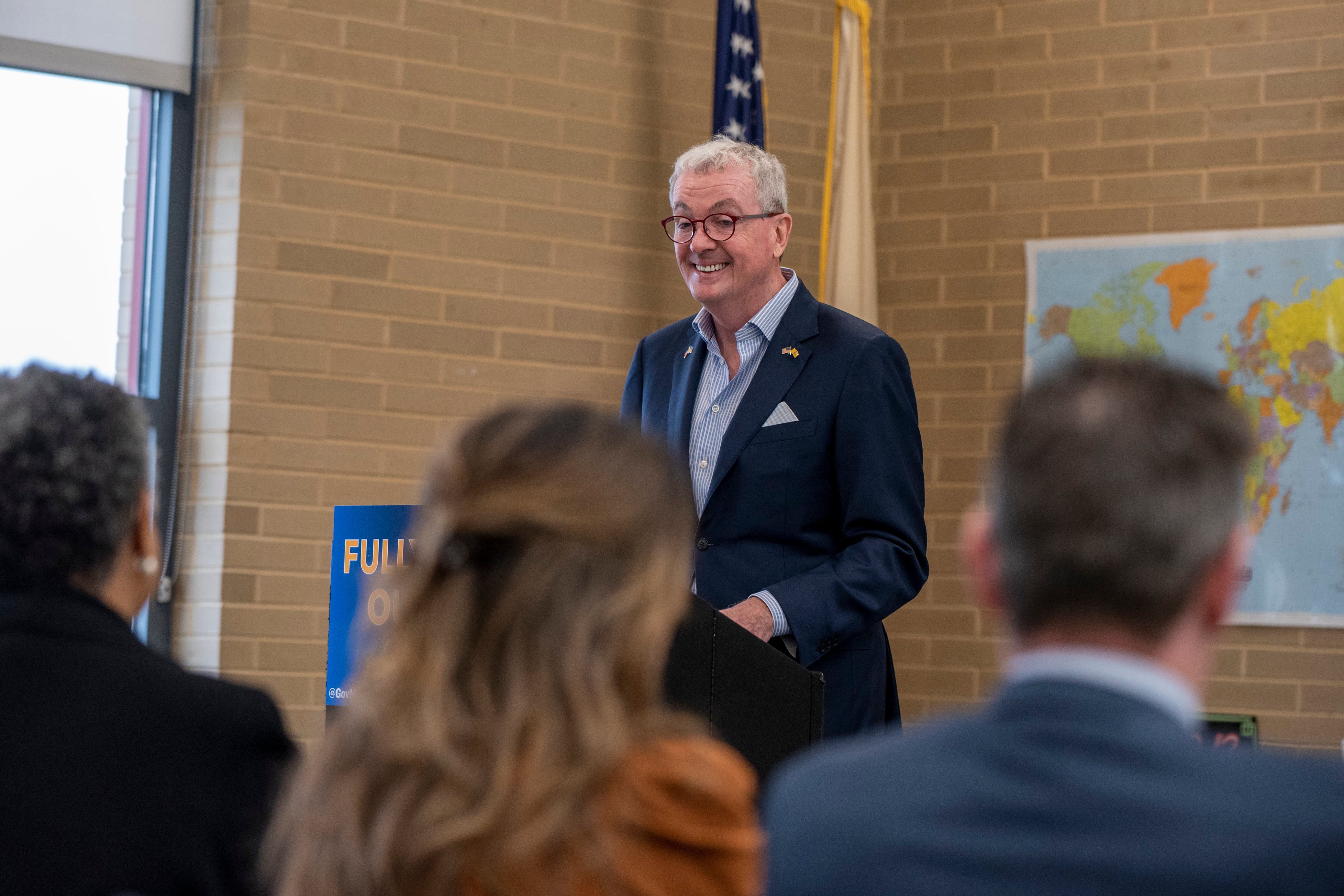 A man in a dark suit jacket in a school building addresses a group of people turned away from the camera.