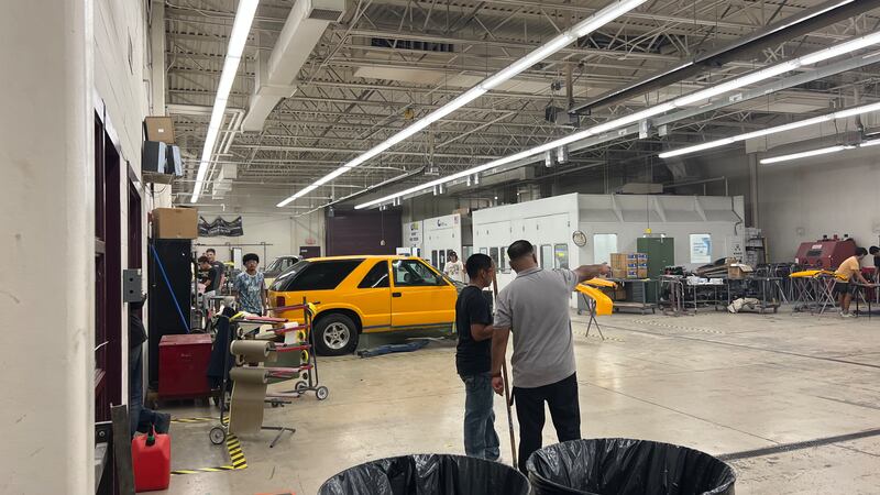 An instructor and student in an automotive garage. There is a yellow SUV in the background.